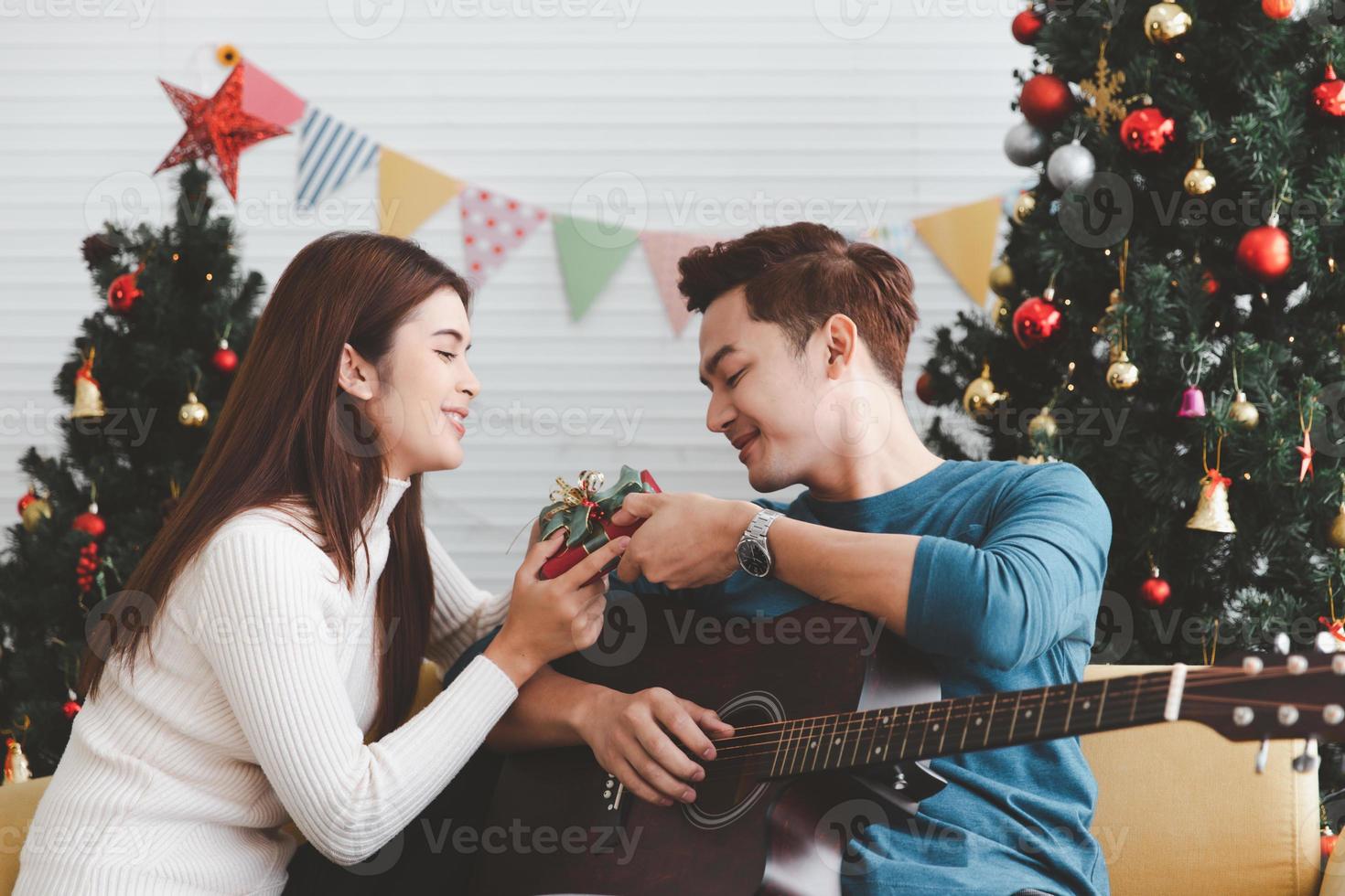 jovem adolescente adulto casal amoroso segurando a caixa de presente e olhando um para o outro durante a celebração do feriado de natal juntos na sala de estar com decoração de enfeite de árvore de natal foto