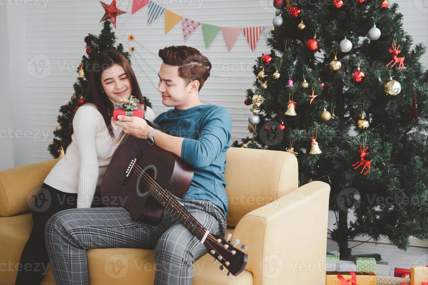 jovem adolescente adulto casal amoroso segurando a caixa de presente e olhando um para o outro durante a celebração do feriado de natal juntos na sala de estar com decoração de enfeite de árvore de natal foto