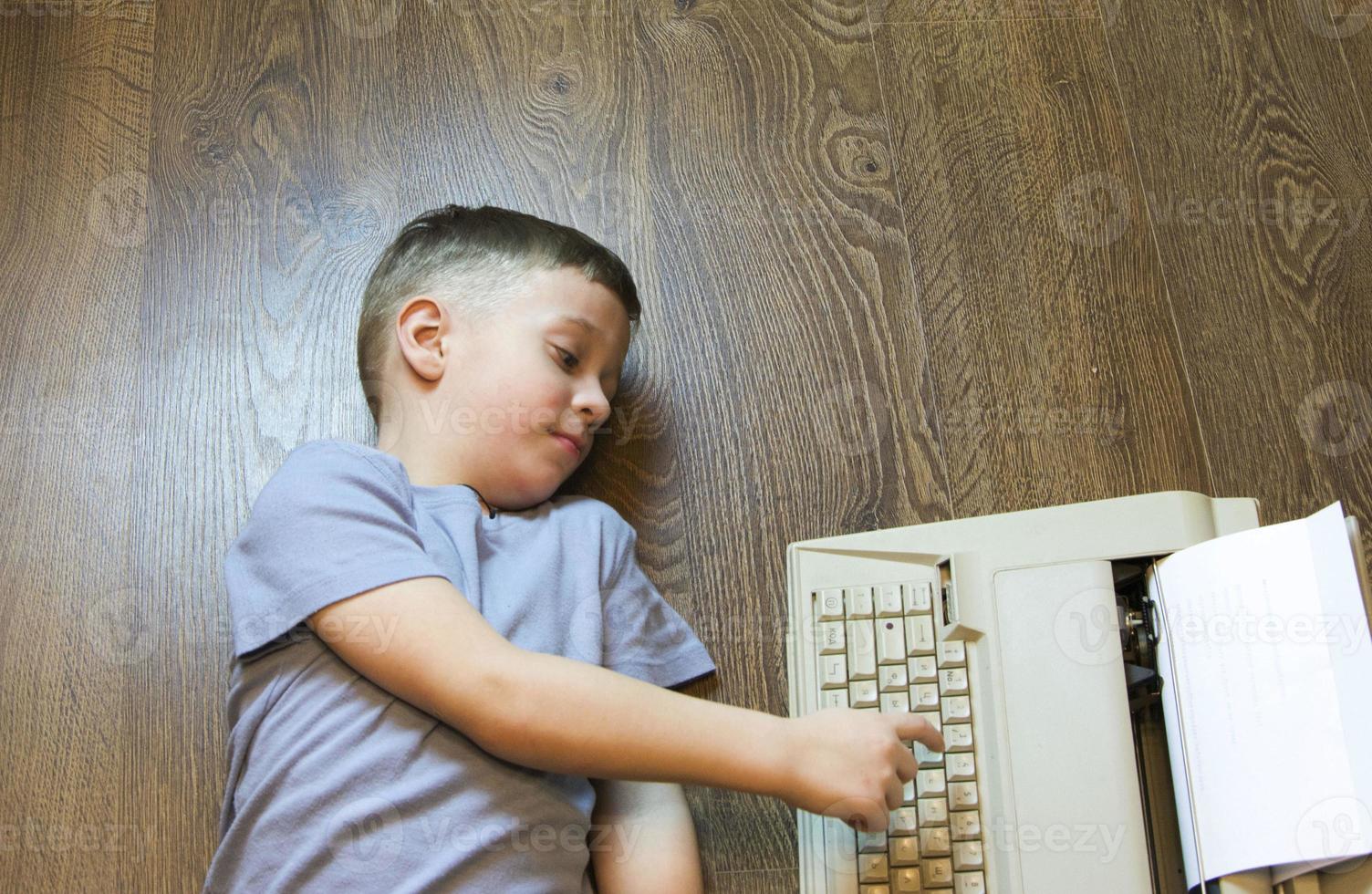 um menino jornalista em idade pré-escolar está deitado no chão e digitando em uma velha máquina de escrever. uma criança usando uma máquina de escrever. jornalista, escritor foto