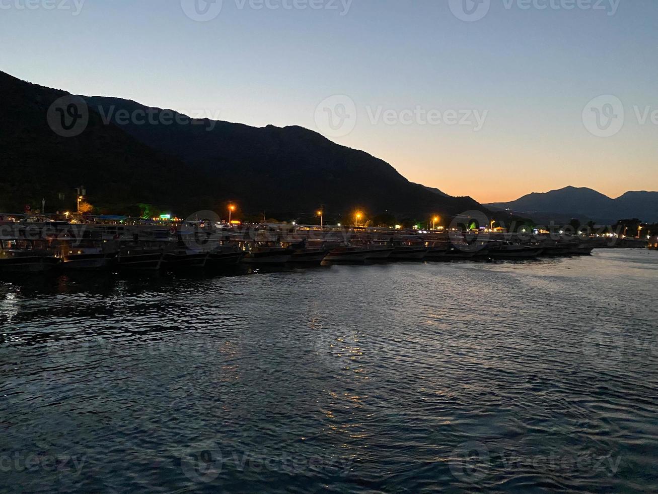 bela costa e praia com contornos de montanhas contra o pano de fundo do pôr do sol do mar ou oceano e luzes ardentes da cidade e porto com navios foto