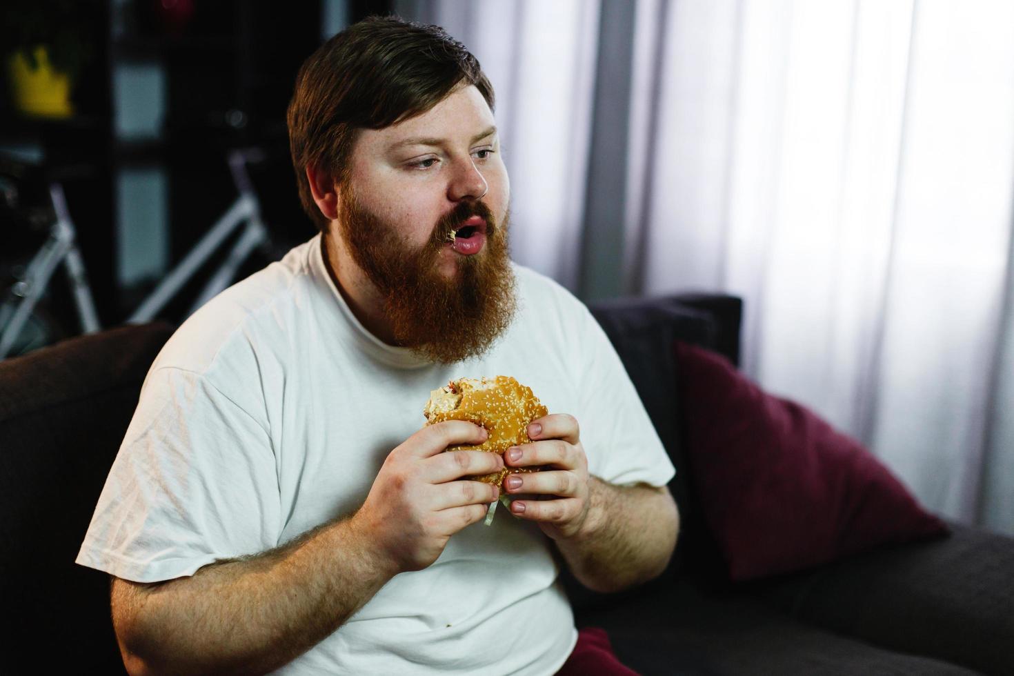 homem gordo sorridente comendo hambúrguer sentado em frente a um aparelho de TV foto