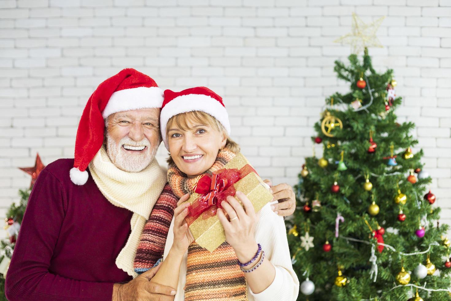 casal caucasiano sorridente feliz comemorando o natal juntos enquanto mostra seu presente de felicidade e emoção em casa com chapéu de papai noel e árvore de natal foto