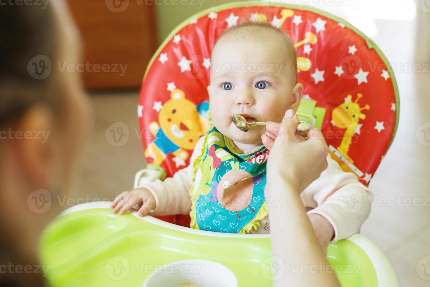 mãe alimenta bebê engraçado de uma colher. criança come em uma cadeira alta foto