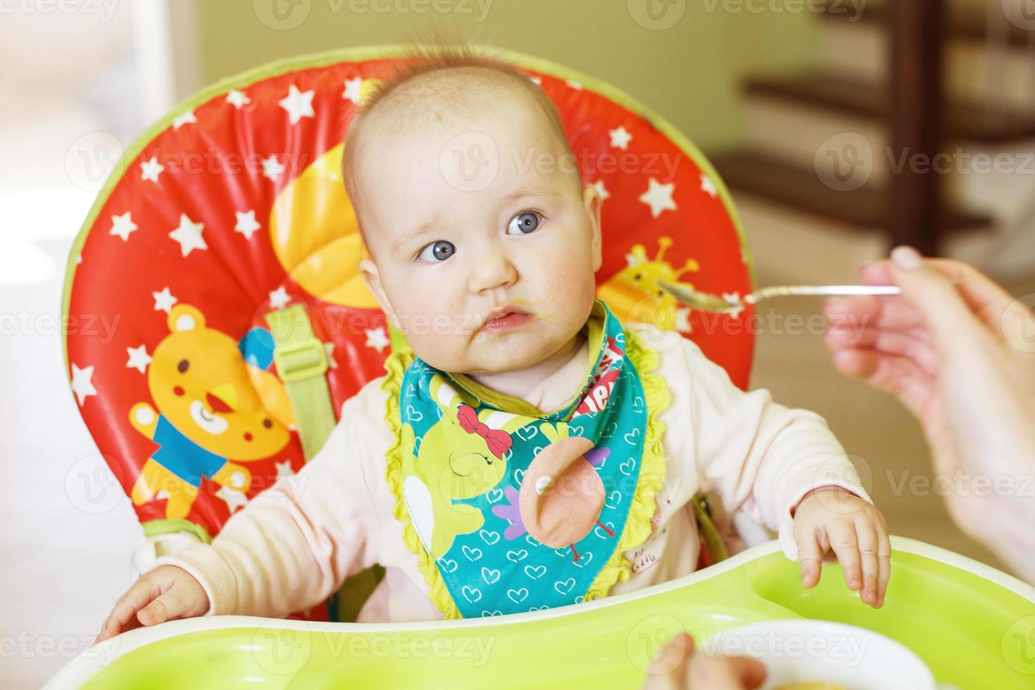 mãe alimenta bebê engraçado de uma colher. criança come em uma cadeira alta foto
