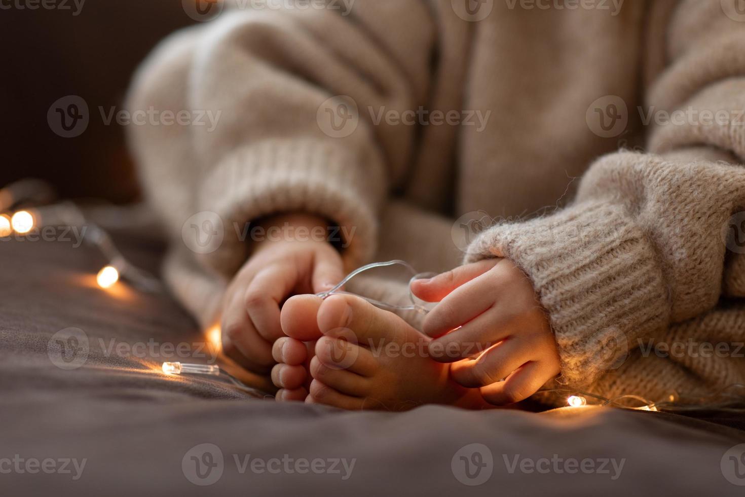 mãos e pés descalços de crianças fecham luzes de guirlanda. suéter bege de malha felpudo felpudo e quente. conceito de natal, feriado. feliz ano novo. foco seletivo. menina criança sentada no sofá dentro de vibrações aconchegantes foto