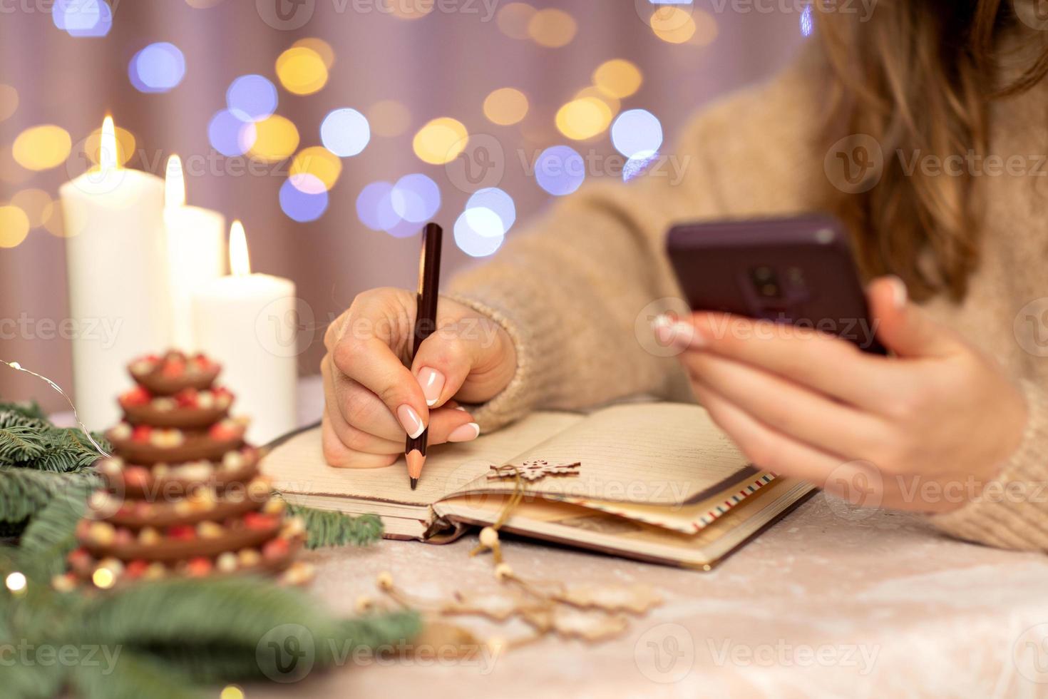 lista de desejos para o conceito de natal de ano novo escrevendo em mãos de notebook.woman segurando uma caneta e um telefone celular. planos de metas. cor de fundo bege. planejar para o próximo ano.online foto