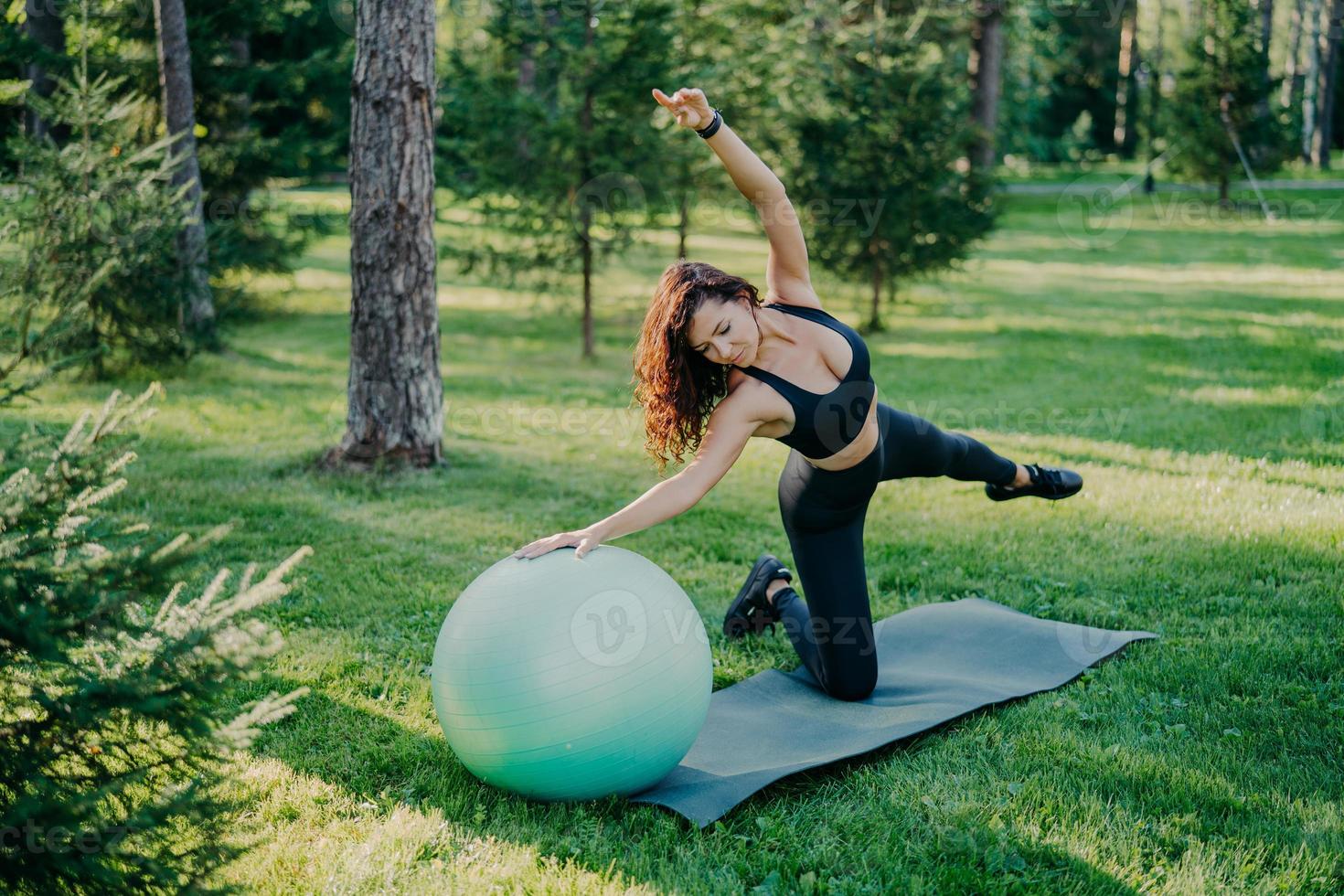 mulher ativa flexível em roupas esportivas faz exercícios de fitness no karemat com fitball, levanta os braços e respira ar fresco na floresta tem poses de treino matinal sobre o fundo da natureza. estilo de vida saudável foto