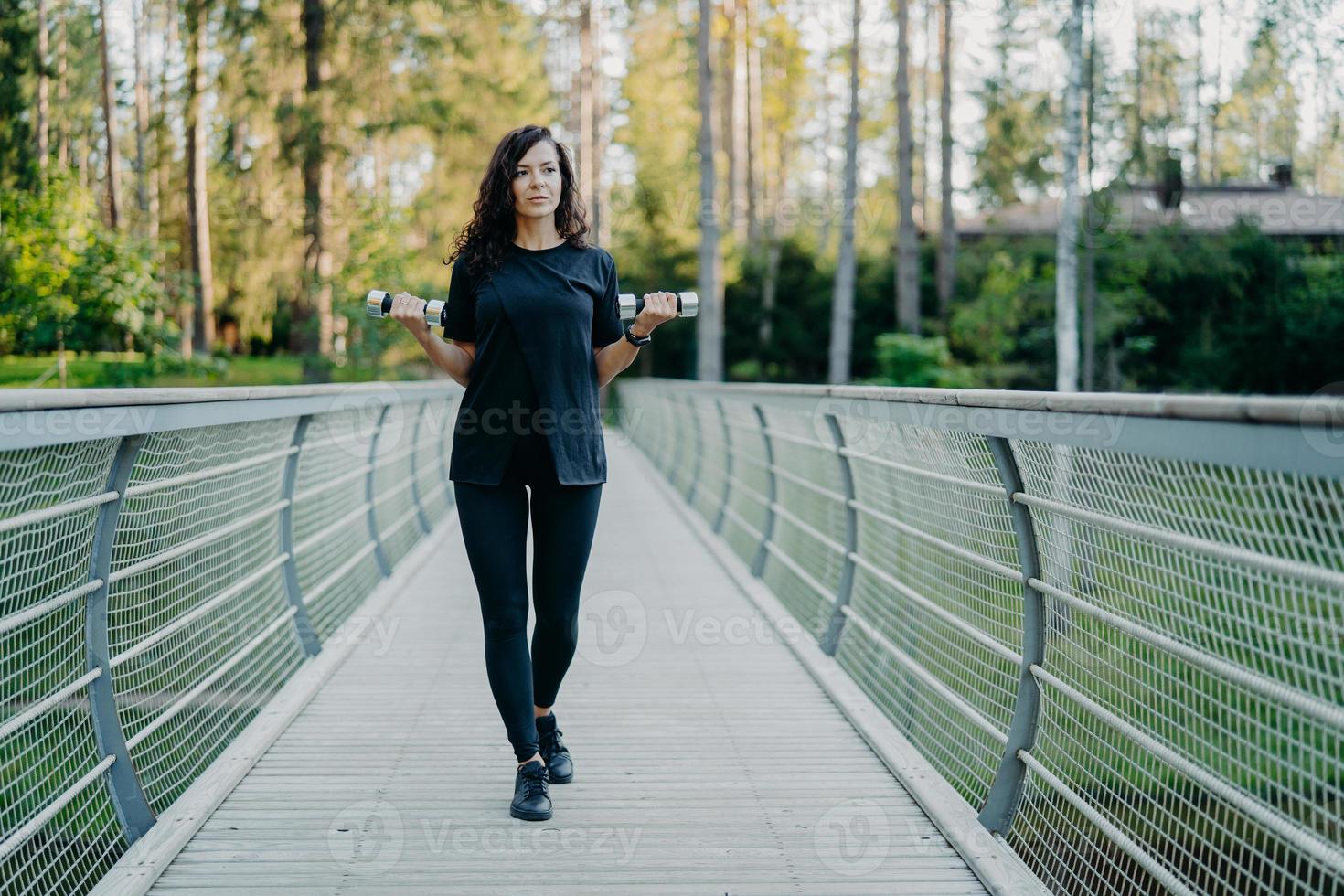 mulher europeia desportiva de camiseta preta e leggings, atravessa a ponte, segura dois halteres, trabalha no treinamento de bíceps, treina ao ar livre, tem expressão pensativa, posa com equipamentos esportivos foto