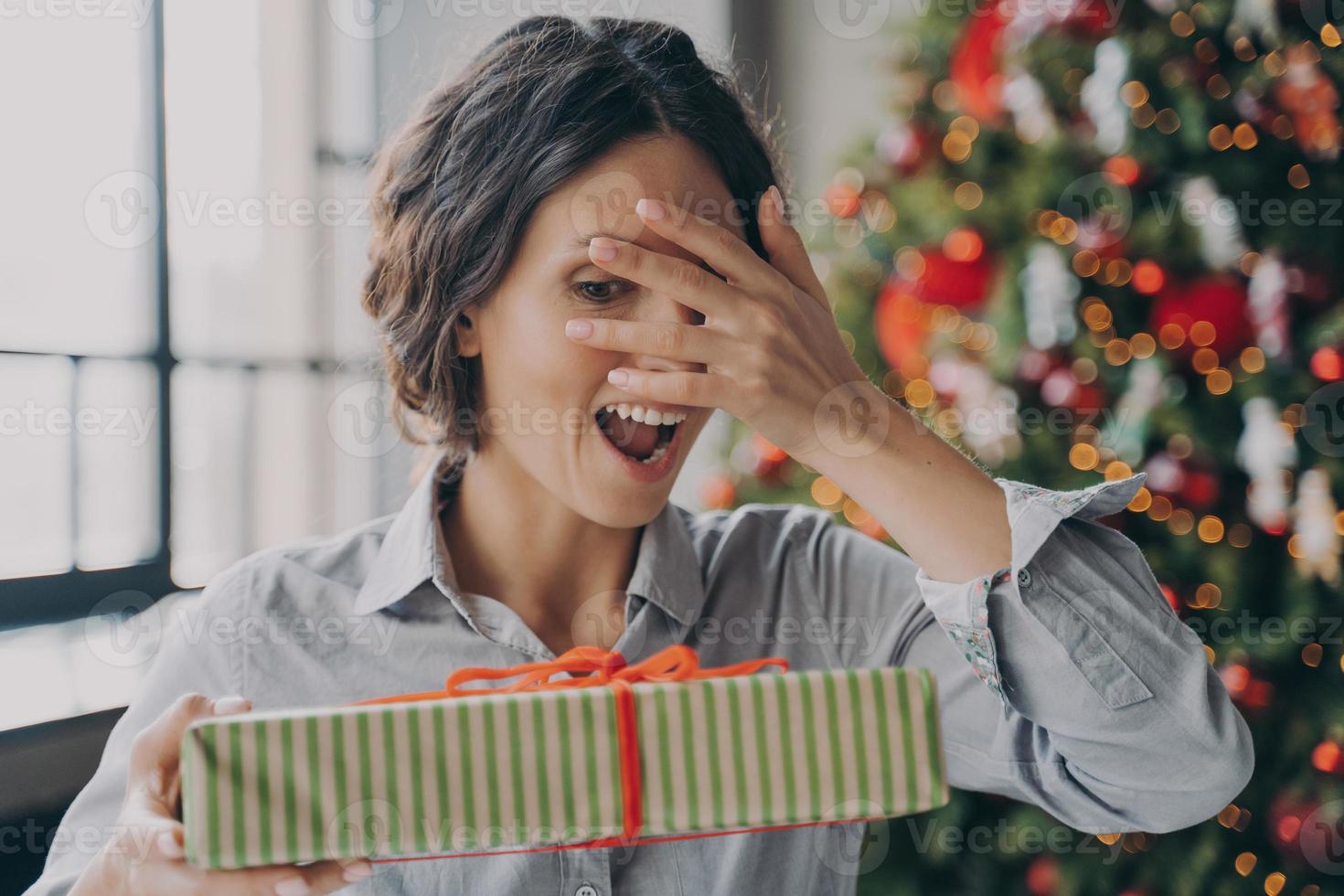 jovem mulher espanhola animada olhando por entre os dedos no presente de natal enquanto comemora o natal em casa foto