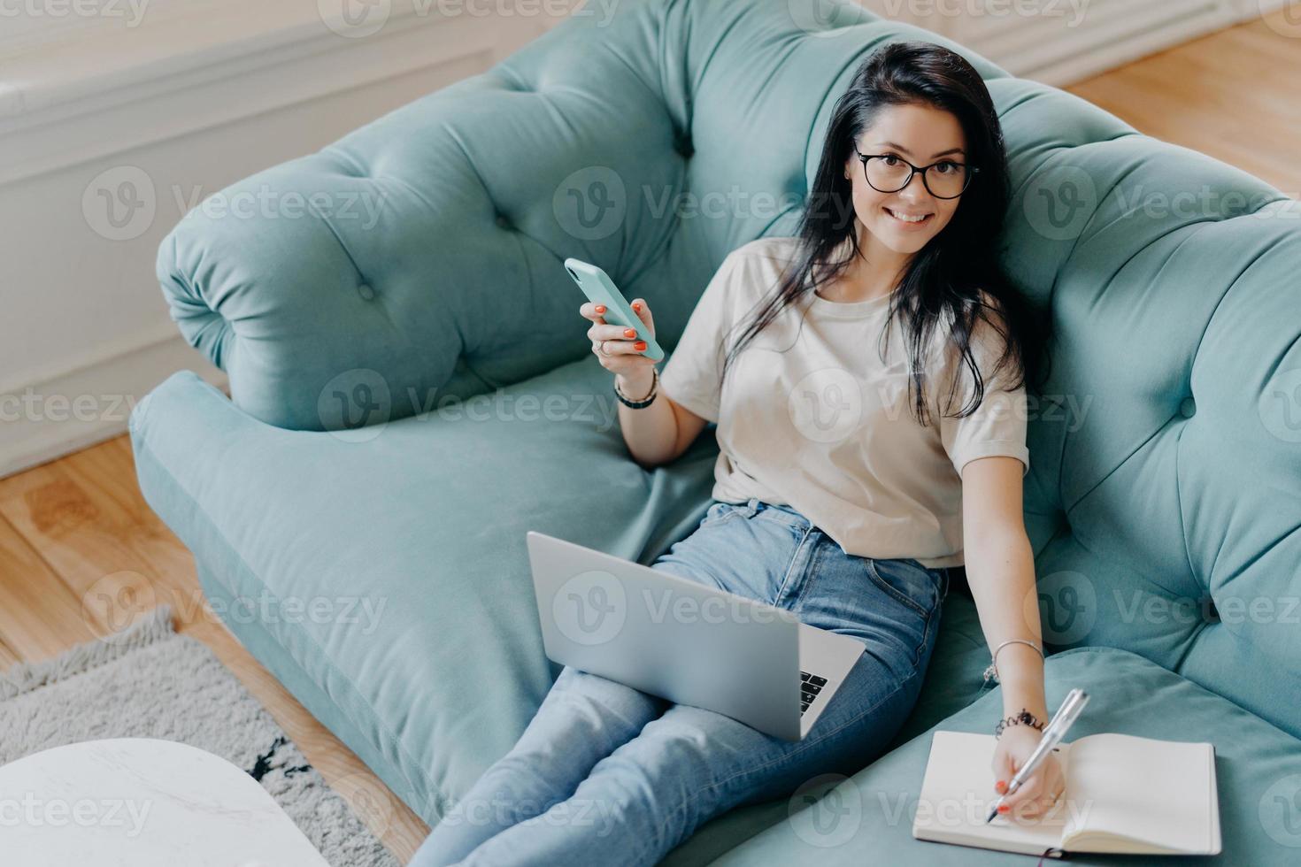 vista de cima do freelancer feminino alegre feliz escreve informações no bloco de notas, usa computador portátil moderno, vestido com roupa casual, senta-se no sofá. estudante prepara trabalho de projeto em casa foto