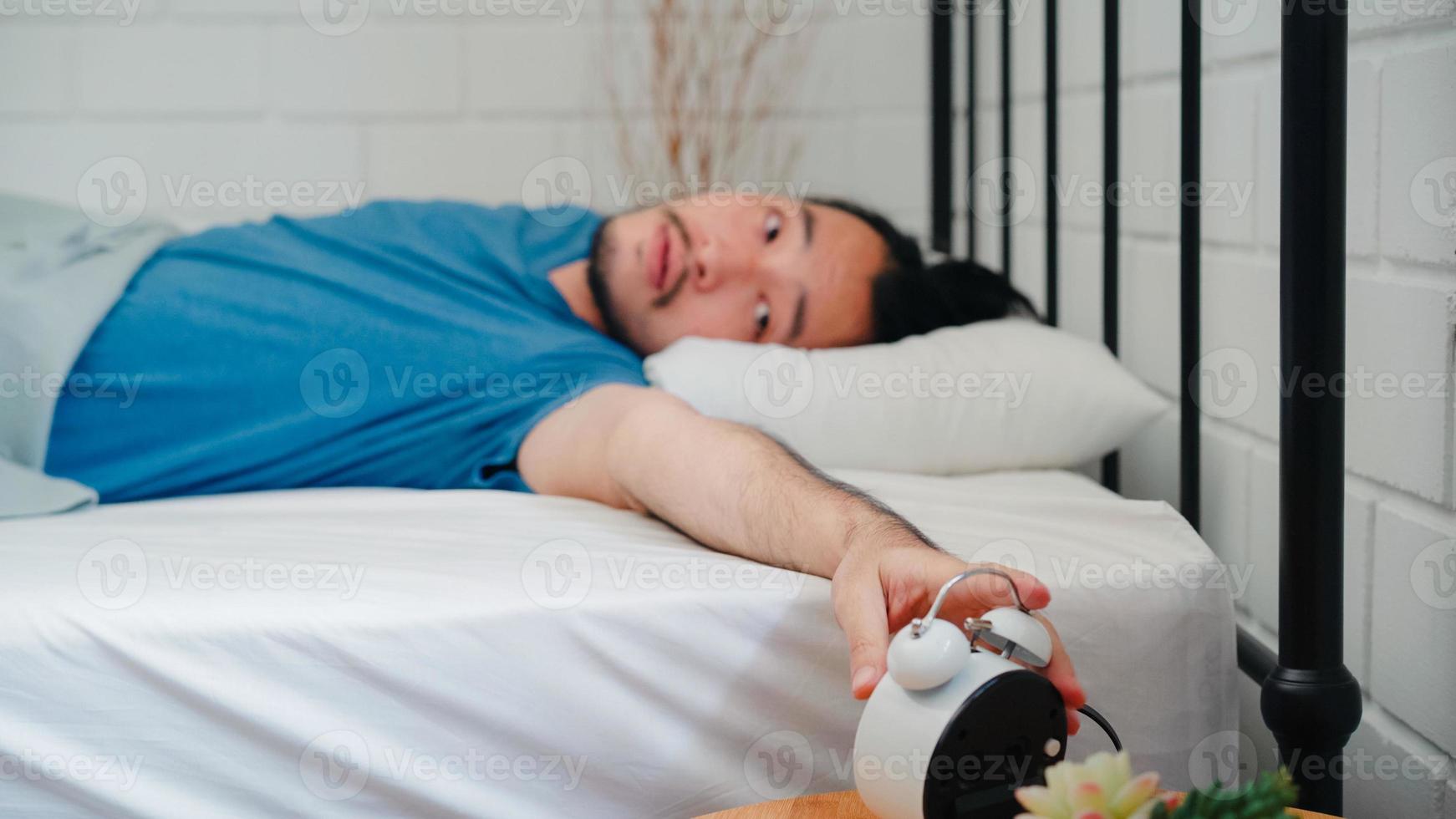 jovem asiático acorda de manhã, homem asiático desliga o despertador depois de acordar na cama no quarto em casa. homens bonitos cochilam, relaxam com sono no conceito de casa moderna. foto