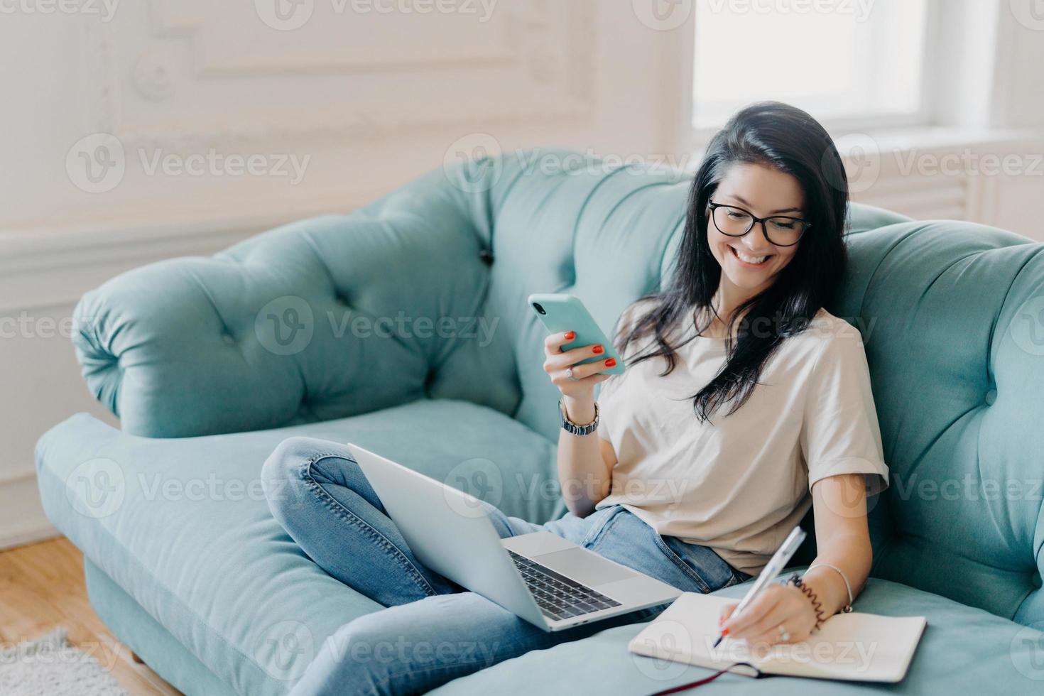 tiro interno de freelancer feminino feliz segura o telefone celular, faz anotações necessárias no diário pessoal, usa tecnologias modernas para compartilhar mídia, senta-se no sofá confortável na espaçosa sala de estar foto