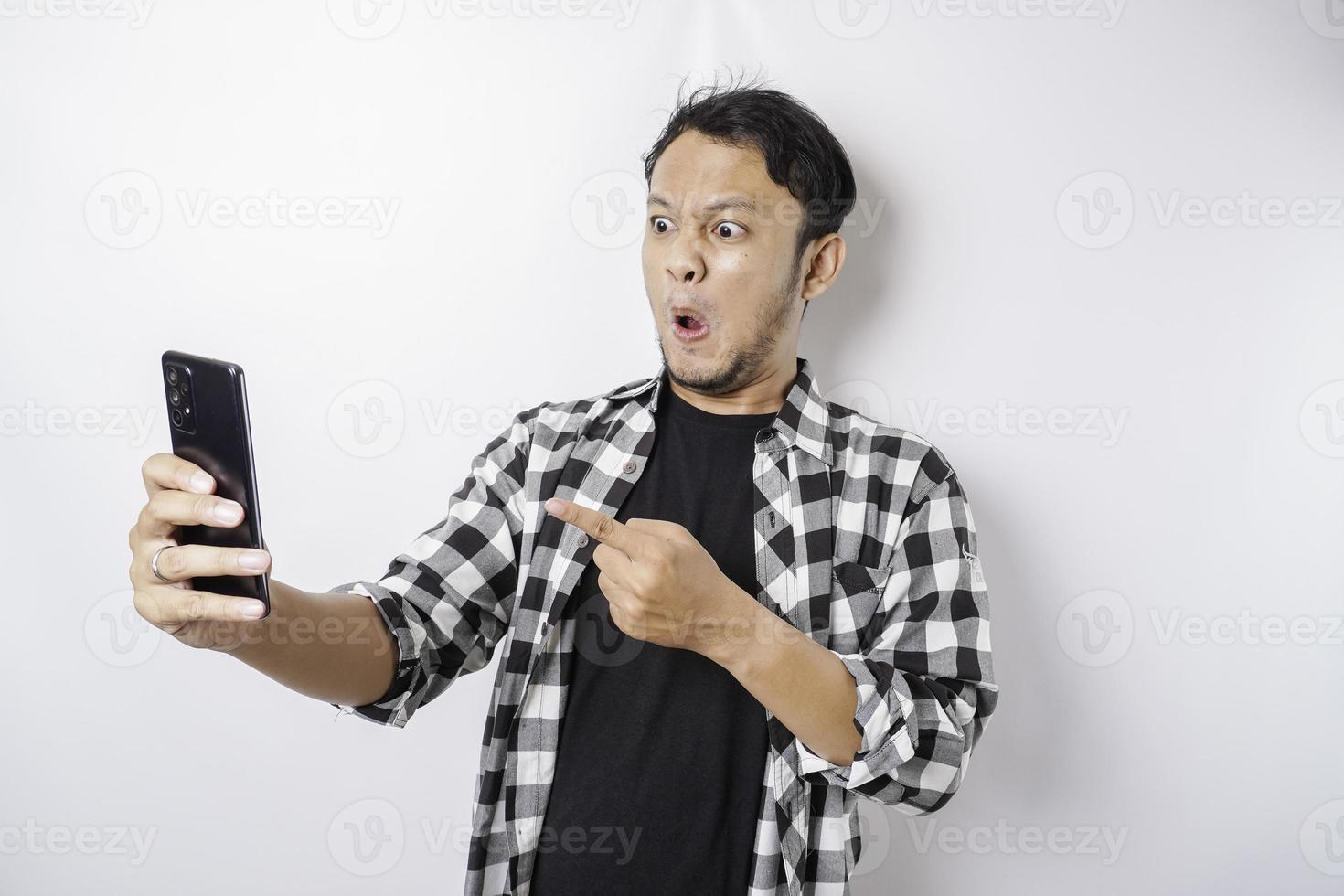 o rosto zangado e louco do homem asiático em camisa xadrez enquanto segura seu telefone no fundo branco isolado. foto