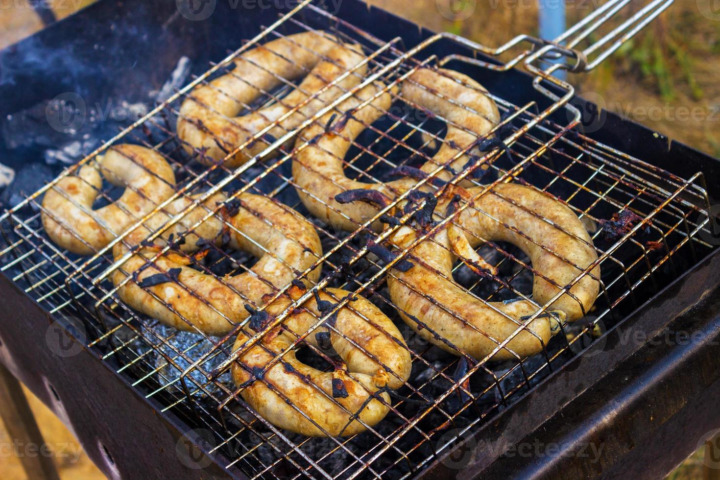 salsichas de porco grelhadas em um churrasco portátil com uma salsicha sendo virada em um par de pinças em um piquenique de verão, close-up da grelha, carne e fogo foto