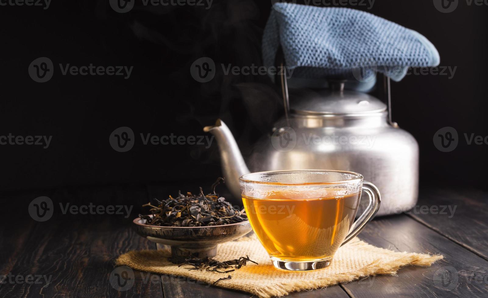 xícara de chá de ervas quente e folhas de chá secas em uma xícara de cerâmica com uma chaleira colocada sobre uma mesa de madeira preta, fundo escuro foto