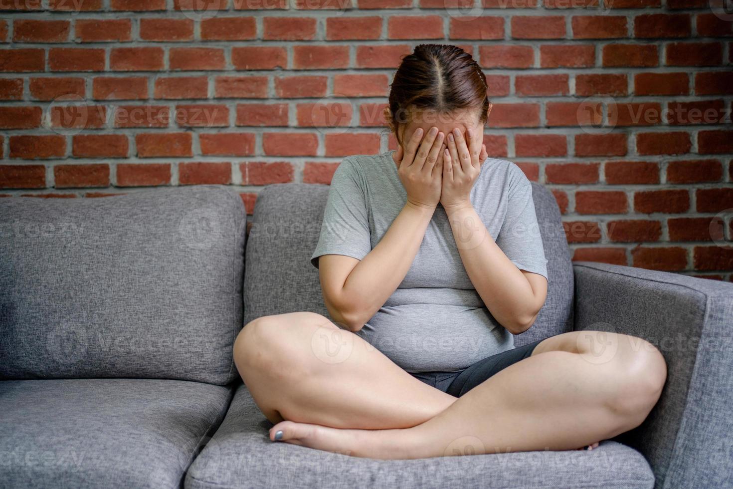 mulher gorda estressada sentada no sofá da sala, sentindo-se desapontada. ela cobriu o rosto com as mãos. foto