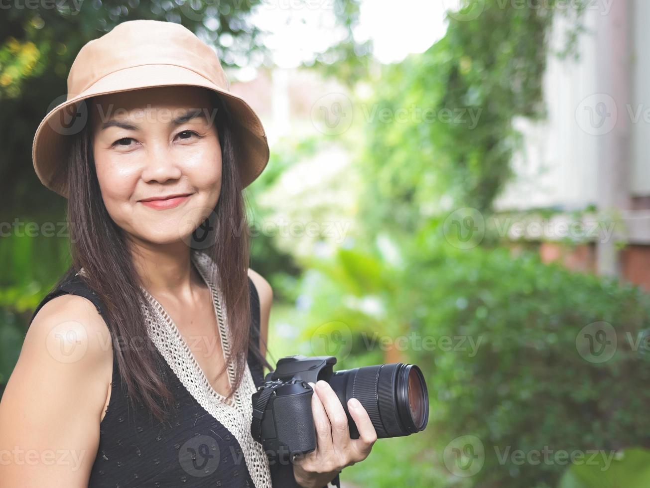 mulher asiática, usando chapéu e top preto sem mangas, de pé no jardim e segurando a câmera dslr, sorrindo alegremente. foto