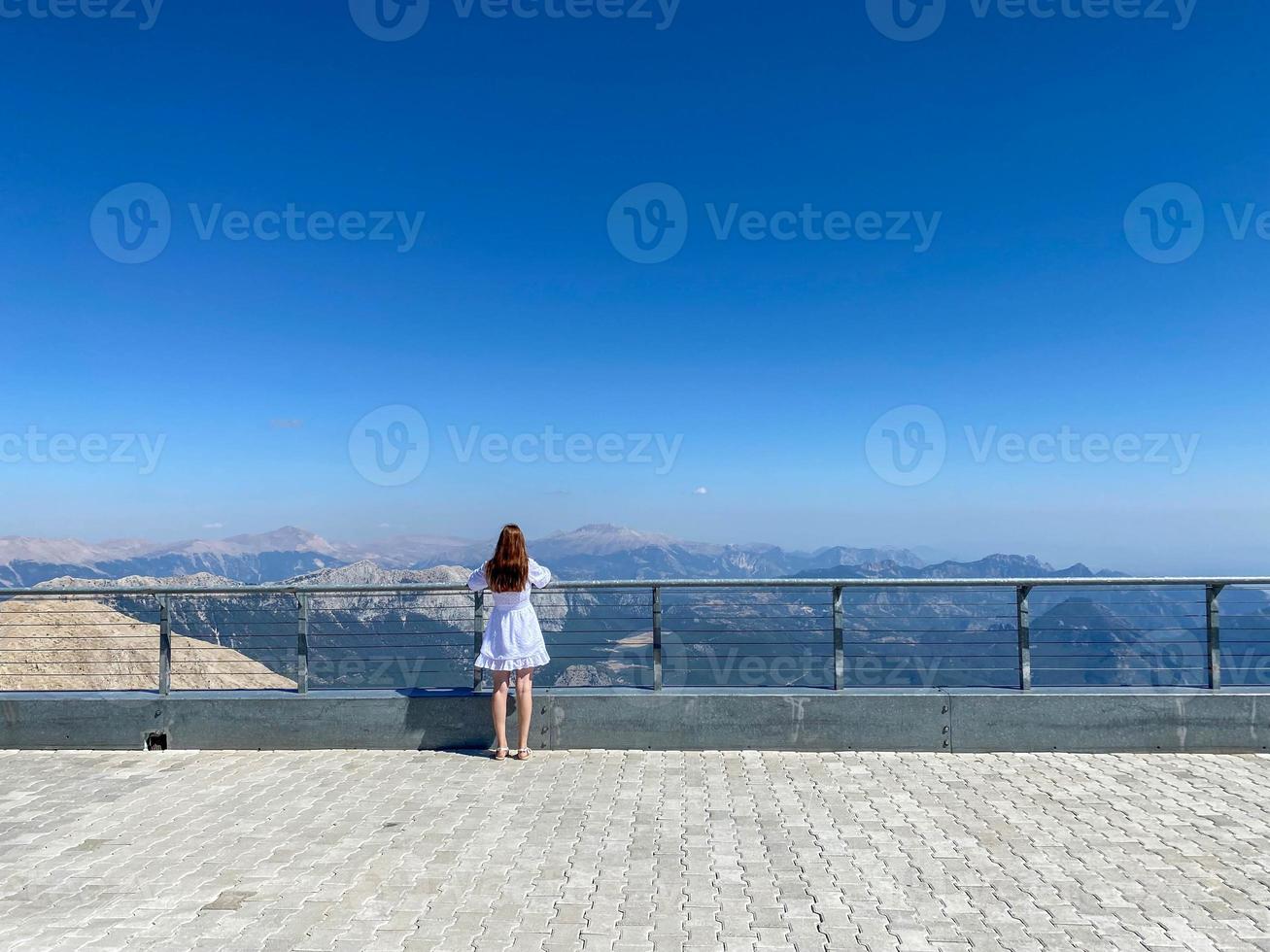 visão traseira do jovem turista feminino asiático em top preto e calças cinza olhando para a câmera em pé ao lado da cerca no deck de observação com colinas verdes e céu azul ao fundo foto