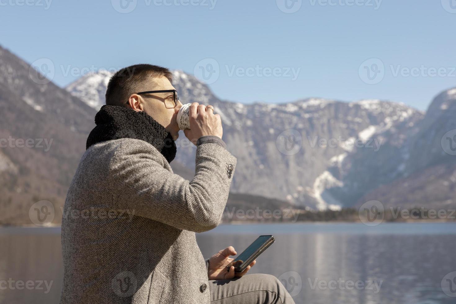 jovem adulto sentado ao ar livre, tomando café e apreciando montanhas, lago, bom tempo, céu azul e sol. paisagem bonita. tempo consigo mesmo, sonho, relaxamento, saúde mental. feriado. foto