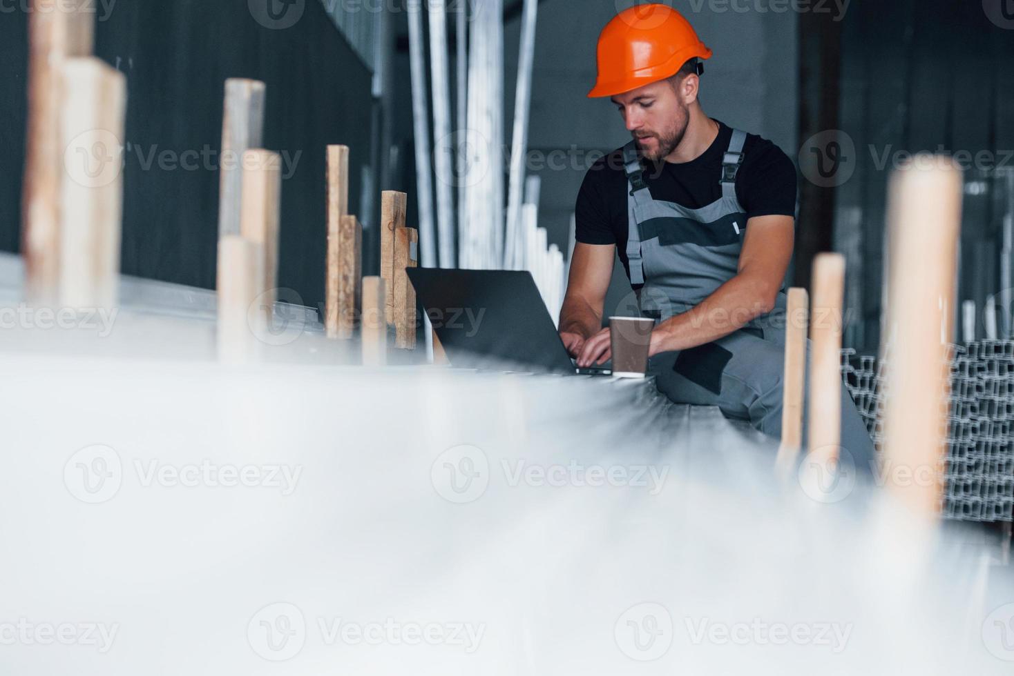 usando laptop de cor prata. trabalhador industrial dentro de casa na fábrica. jovem técnico com capacete laranja foto