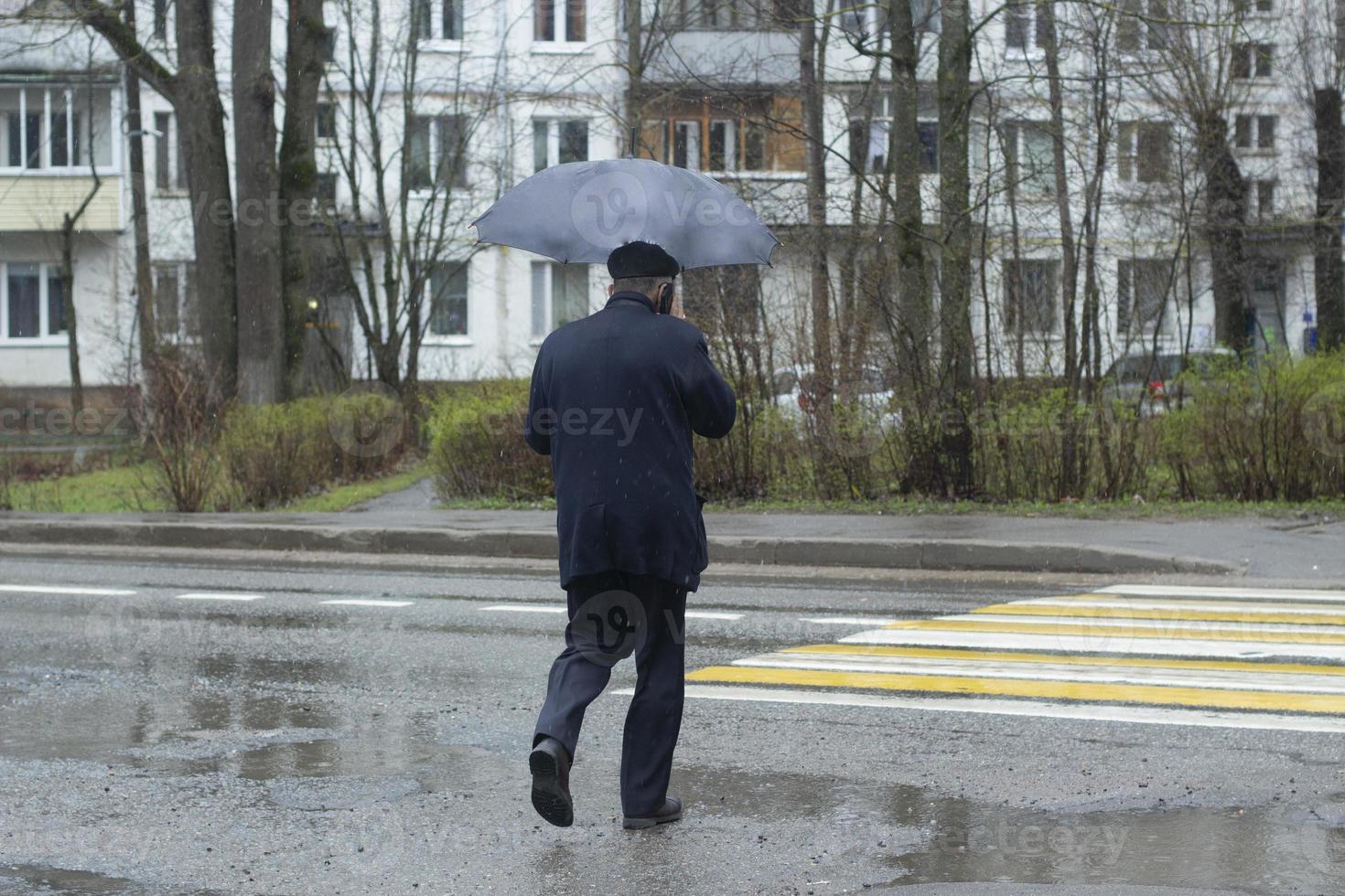 um homem idoso de terno azul com guarda-chuva atravessa a rua em uma passagem para pedestres, vista de trás. um dia chuvoso. tráfego. foto