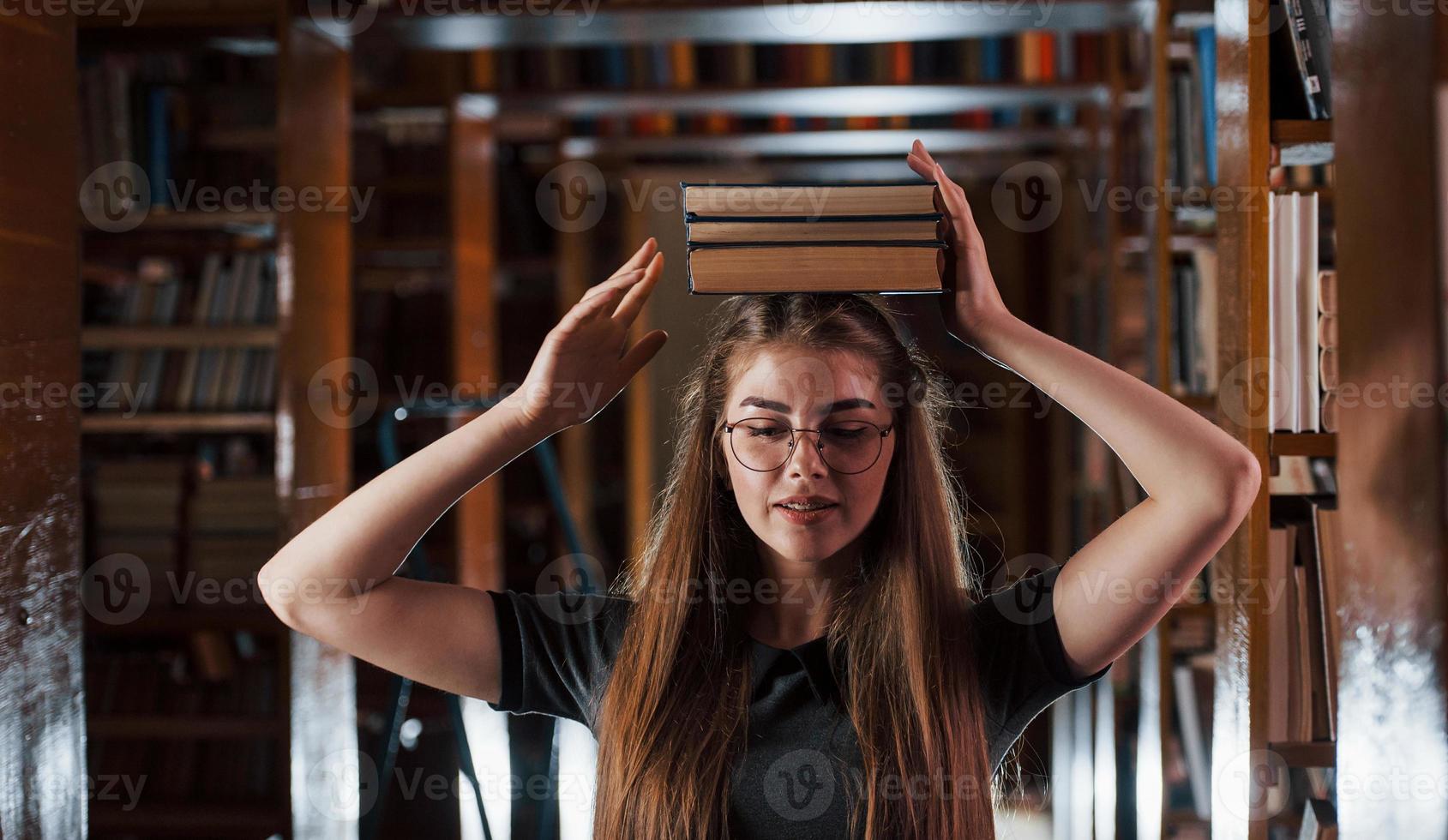 garota alegre se diverte enquanto coloca livros na cabeça. aluna está na biblioteca. concepção de educação foto