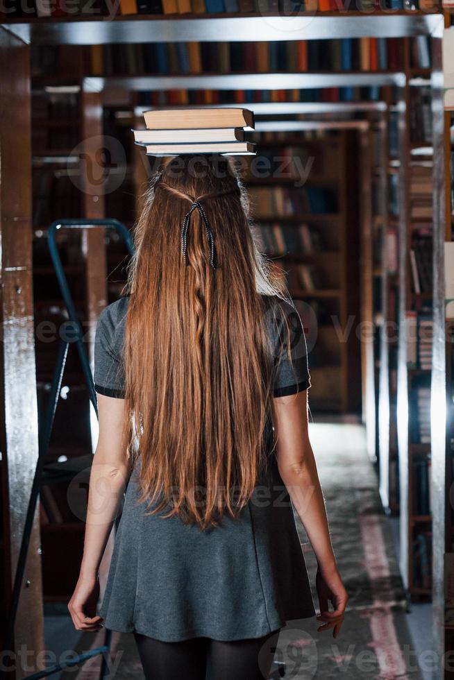 garota alegre se diverte enquanto coloca livros na cabeça. aluna está na biblioteca. concepção de educação foto