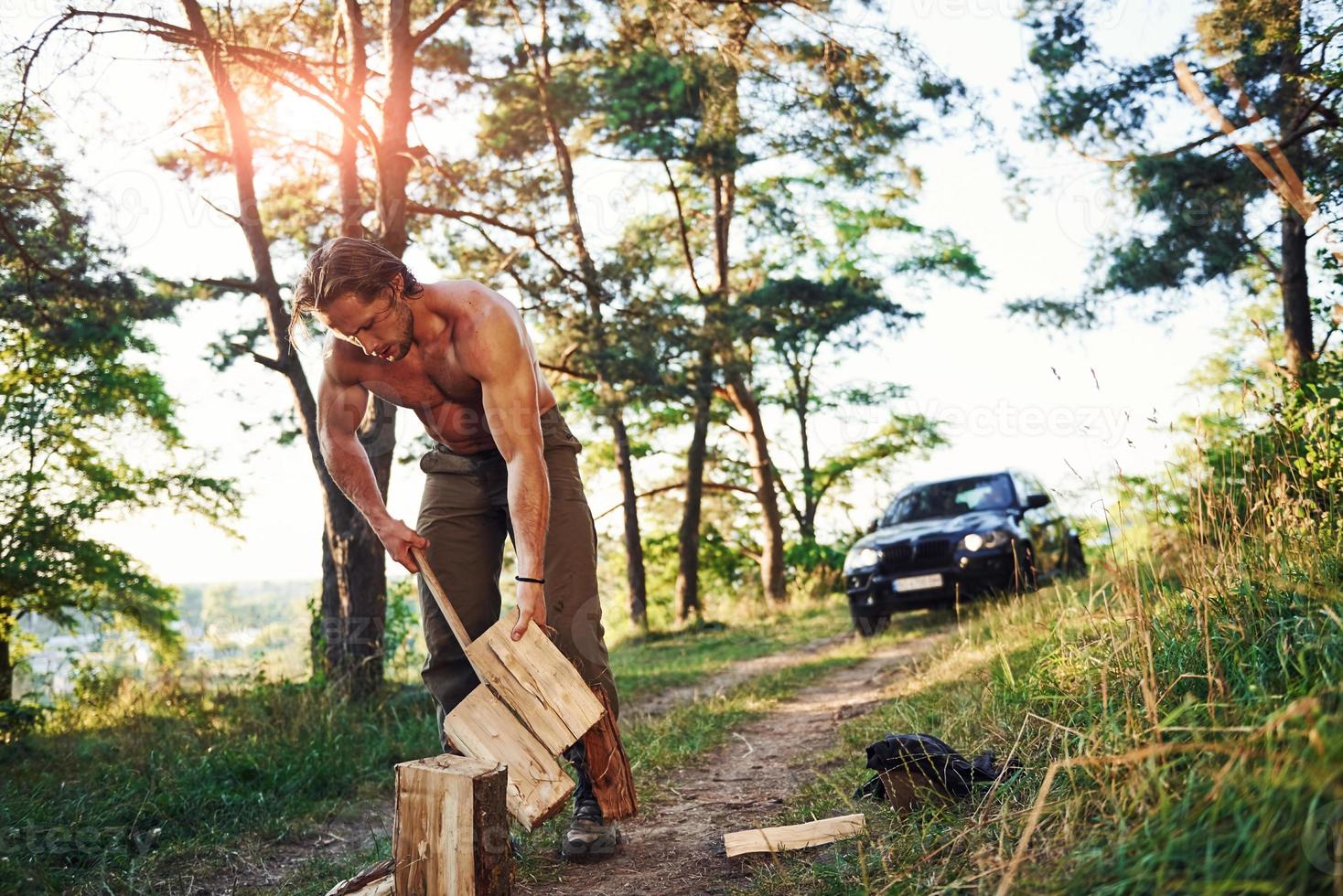 lenhador com um machado cortando madeira. homem bonito sem camisa com tipo de corpo musculoso está na floresta durante o dia foto