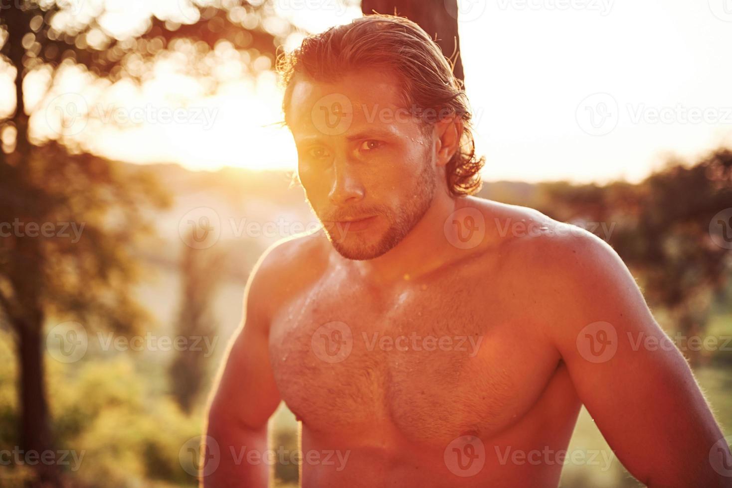 olhar sério e confiante. homem bonito sem camisa com tipo de corpo musculoso está na floresta durante o dia foto