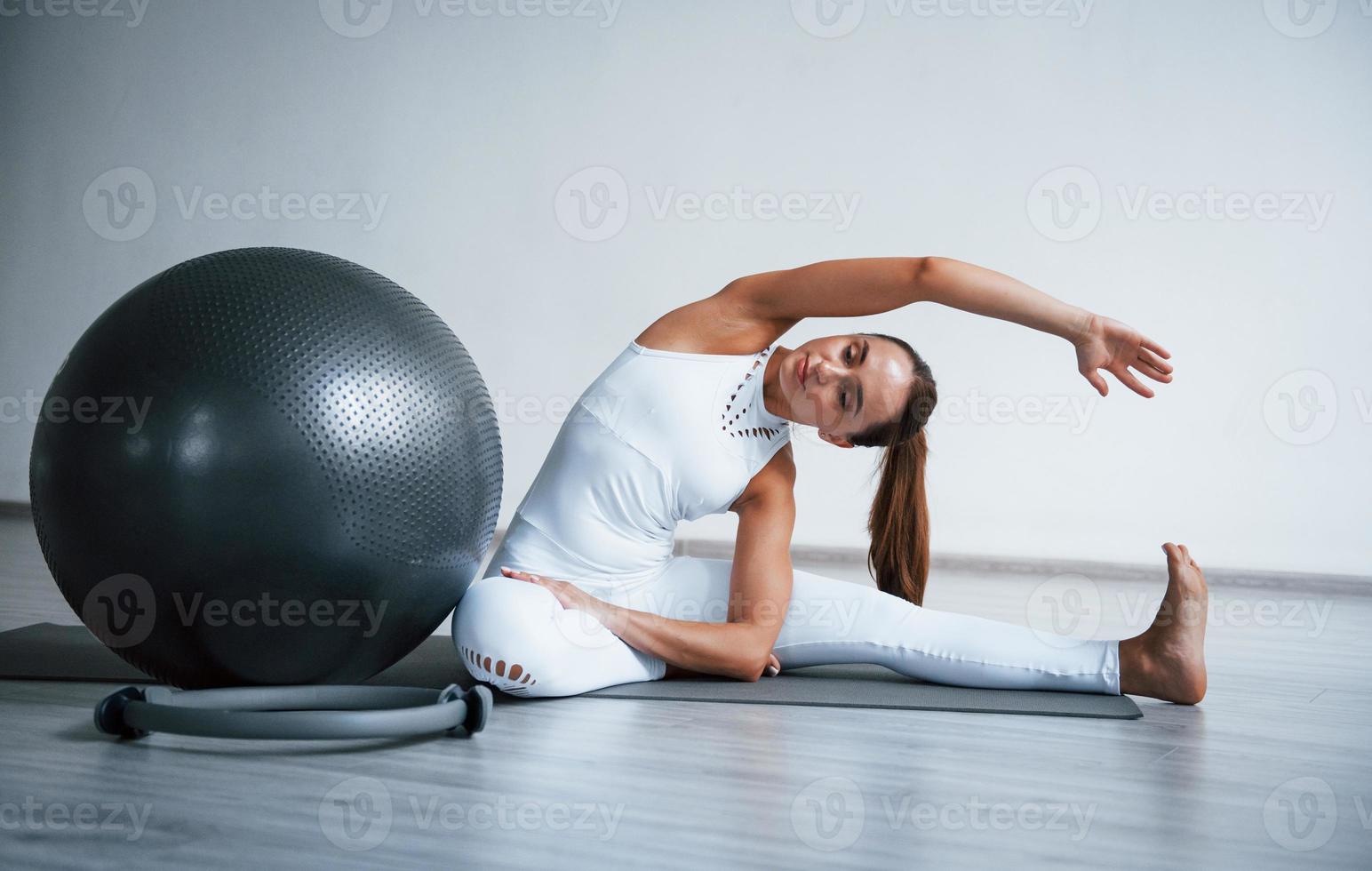 sentado no tapete de rolo. jovem mulher desportiva em roupas brancas com tipo de corpo magro no centro saudável fazendo exercícios. bola de ginástica preta foto