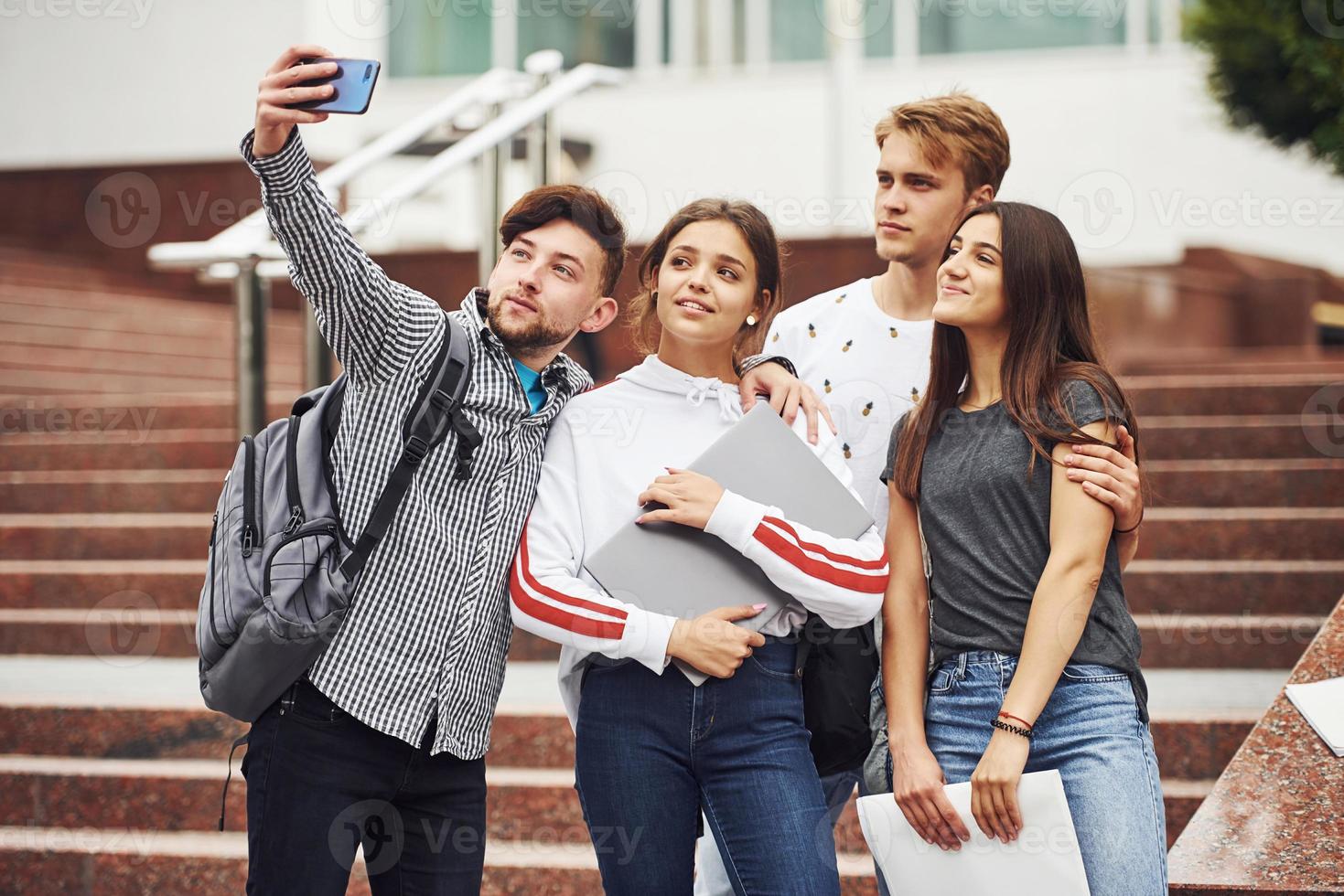 de pé e tirando selfie. grupo de jovens estudantes em roupas casuais perto da universidade durante o dia foto