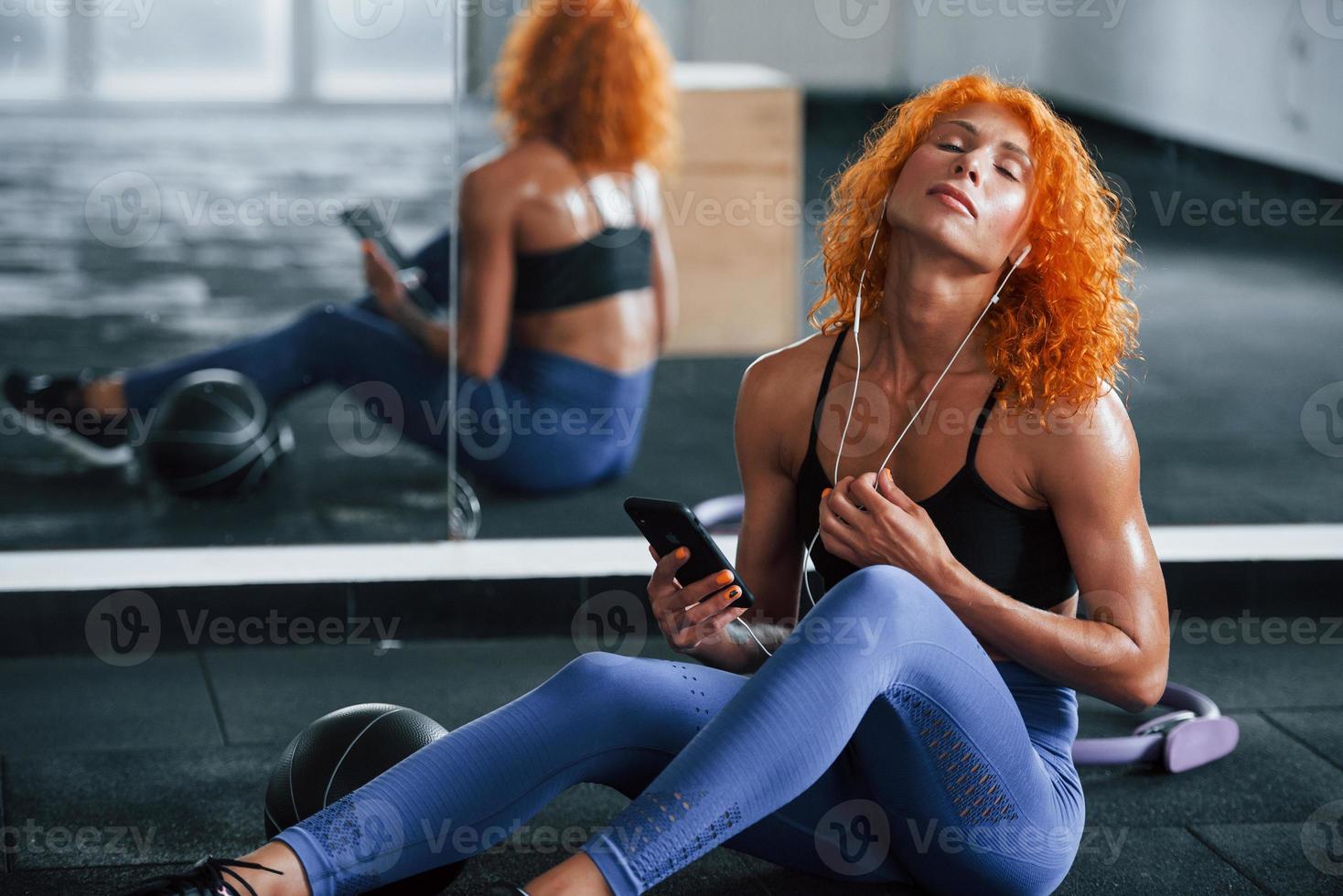 ouvindo a música em fones de ouvido. menina ruiva desportiva tem dia de fitness no ginásio durante o dia. tipo de corpo musculoso foto