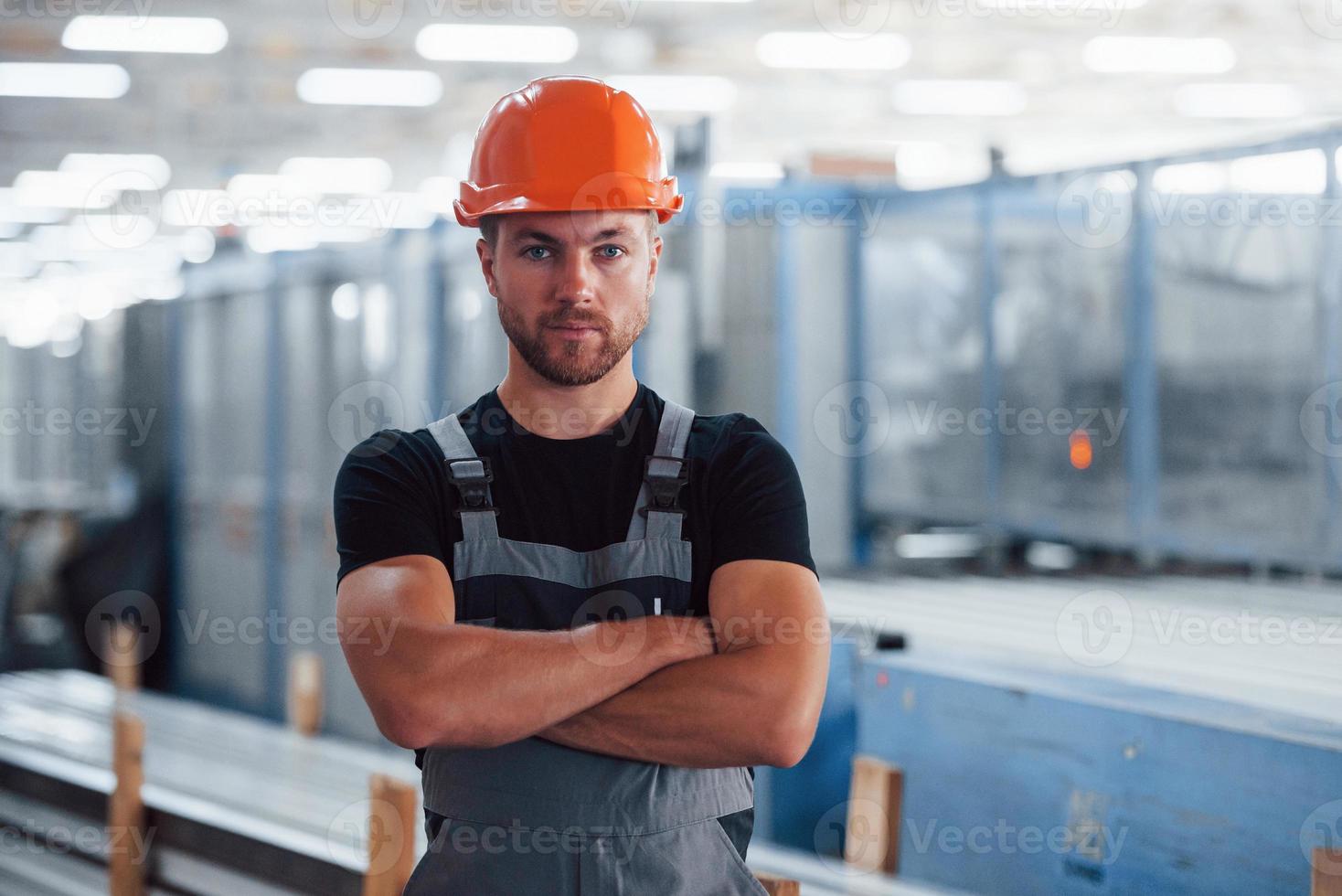 fica de braços cruzados. retrato do trabalhador industrial masculino dentro de casa na fábrica. jovem técnico com capacete laranja foto