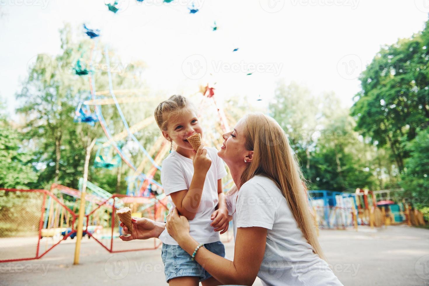 tomando sorvete. menina alegre sua mãe se diverte no parque juntos perto de atrações foto