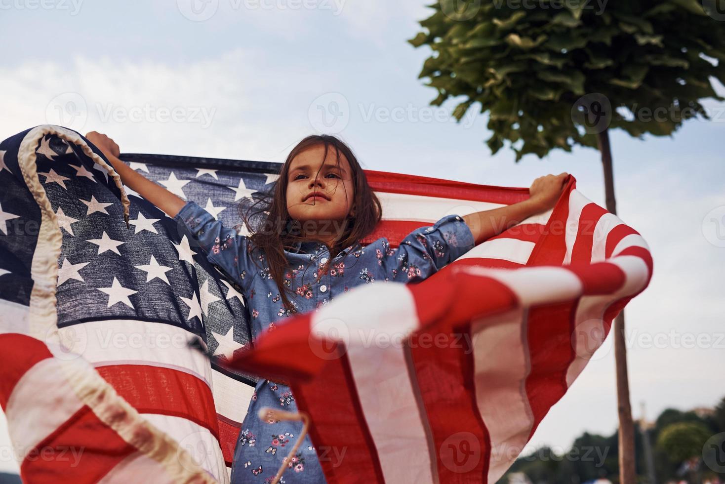 linda árvore verde. criança feminina patriótica com bandeira americana nas mãos. contra o céu nublado foto