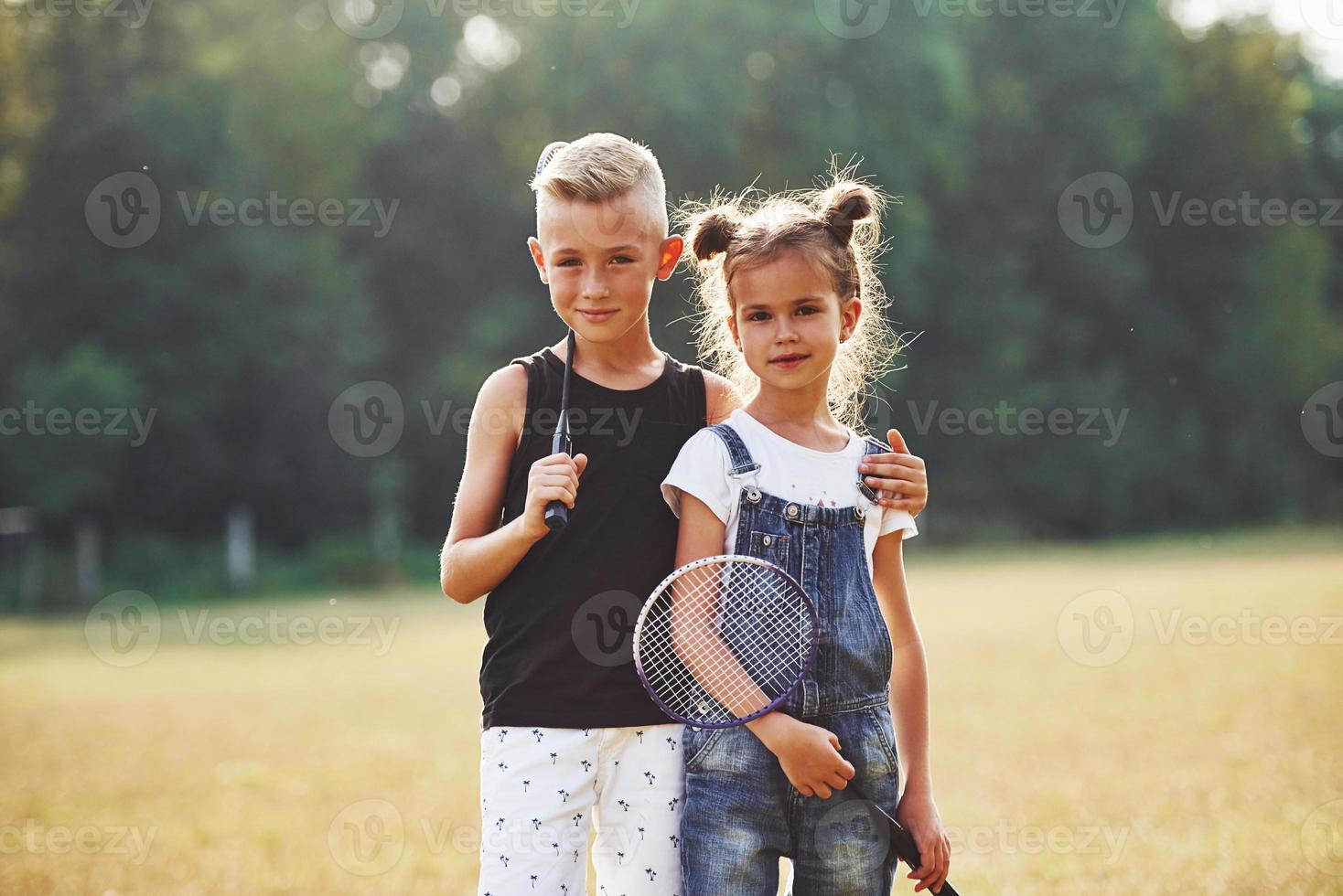 retrato de menino e menina em pé no campo em dia ensolarado com raquetes de tênis nas mãos foto
