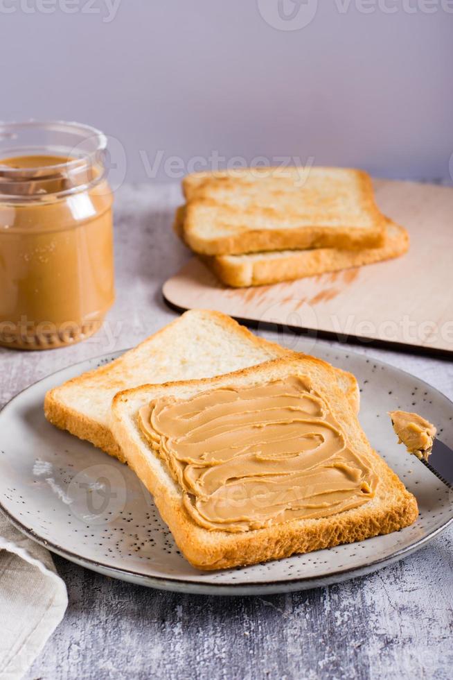 sanduíche de manteiga de amendoim no pão torrado em um prato e pote de manteiga na mesa. visão vertical foto