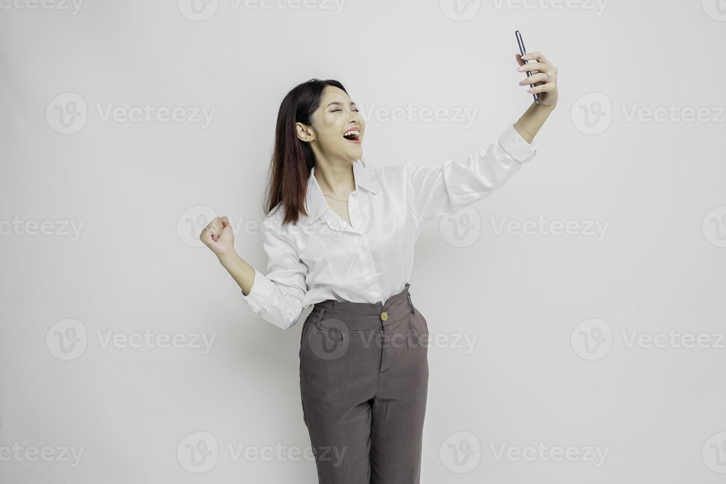 uma jovem mulher asiática com uma expressão de sucesso feliz vestindo camisa branca e segurando o smartphone isolado pelo fundo branco foto