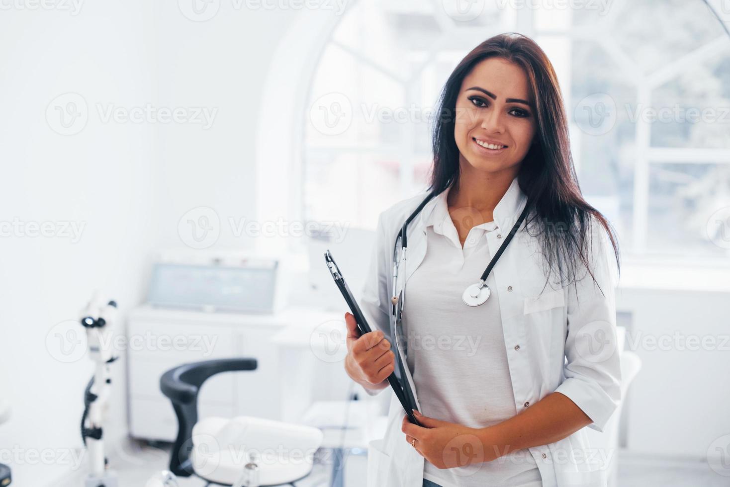 com o bloco de notas nas mãos. médica morena fica no escritório moderno e olha para a câmera foto