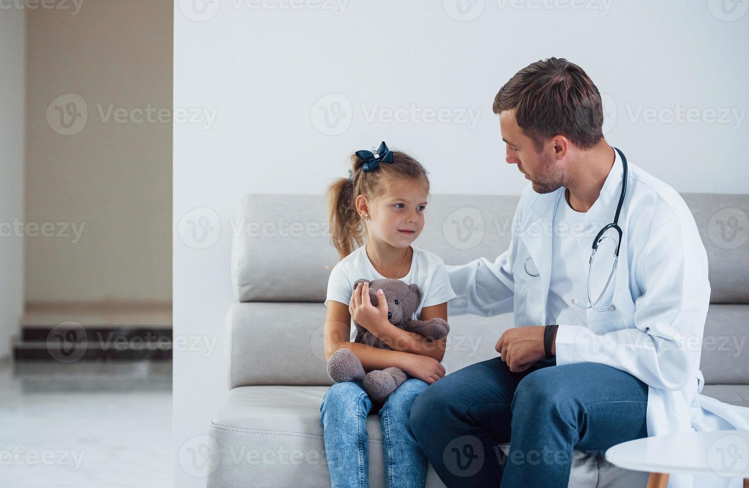 médico masculino de uniforme branco senta-se na clínica com a garotinha foto