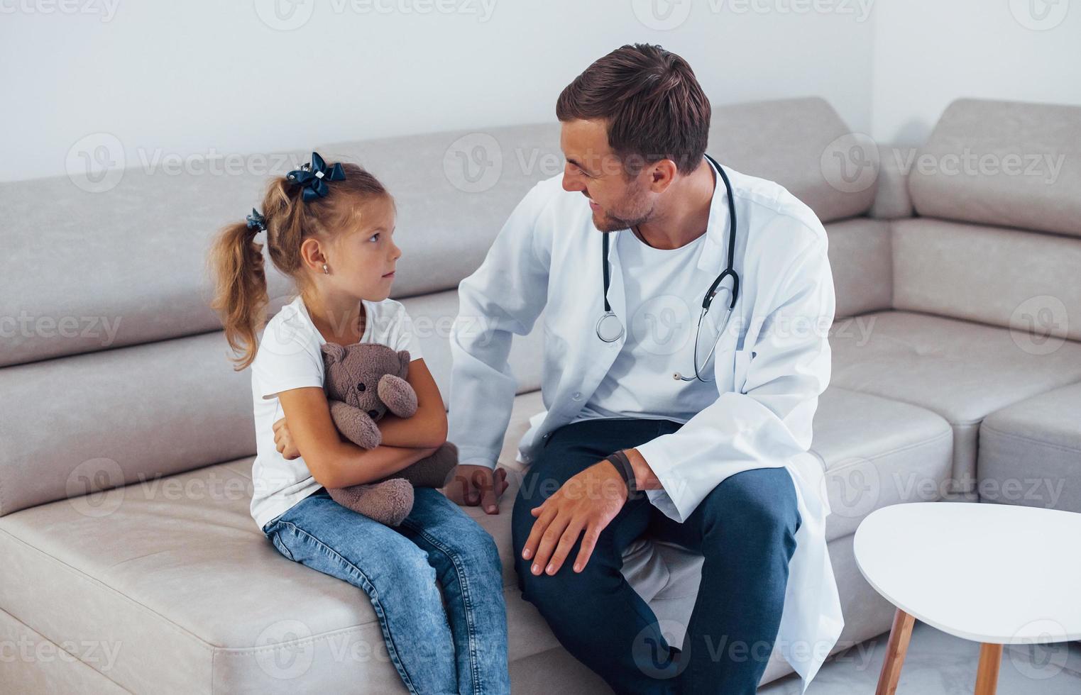 médico masculino de uniforme branco senta-se na clínica com a garotinha foto