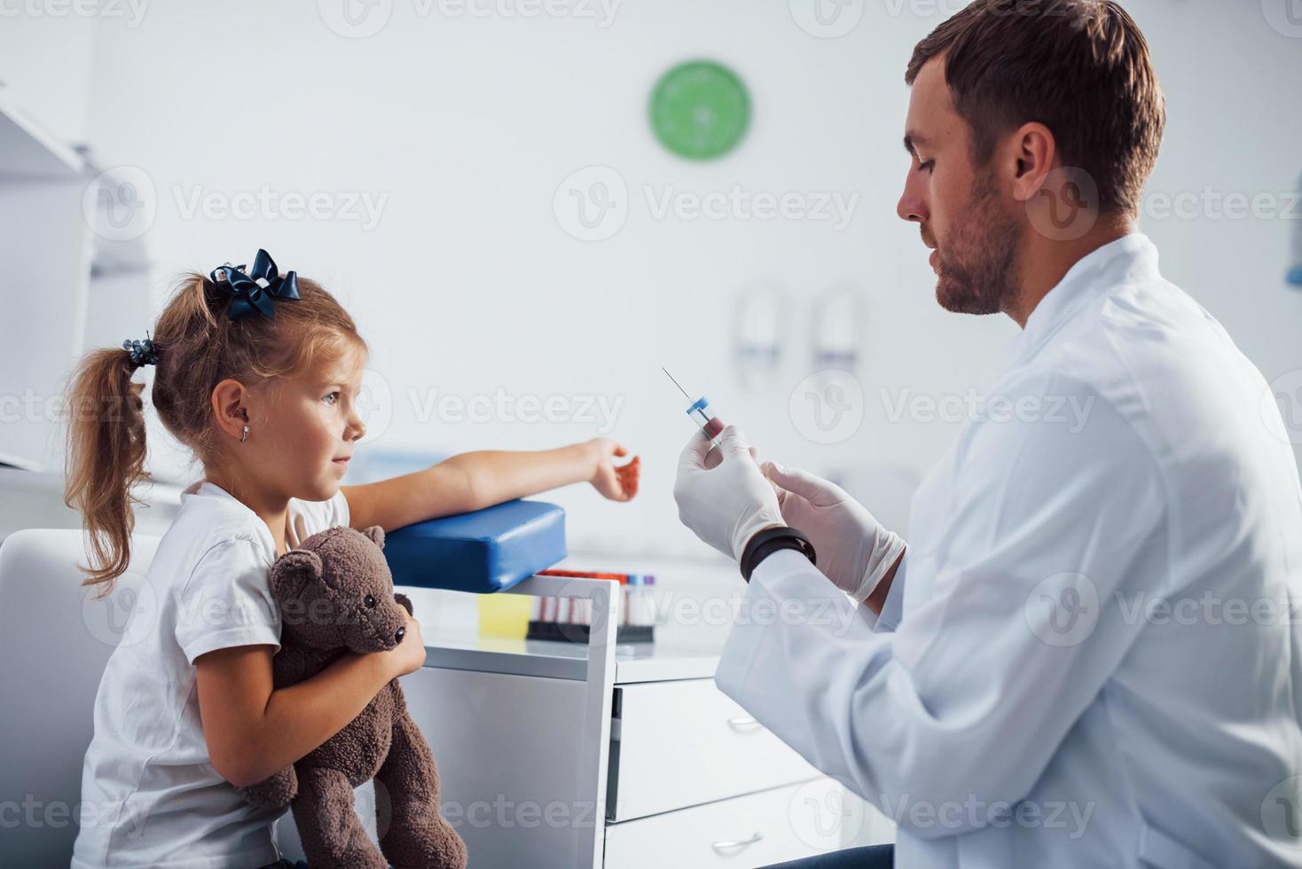 amostra de sangue. menina com seu ursinho de pelúcia está na clínica com o médico foto