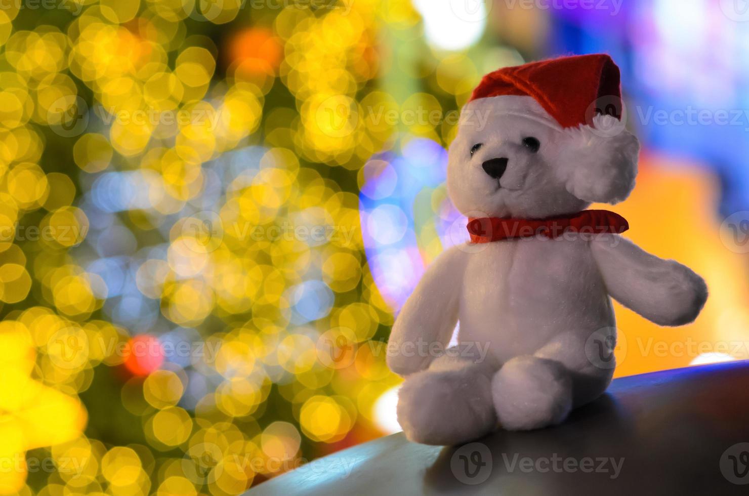 papai noel glitter ursinho branco usando chapéu sentado na frente de luzes coloridas bokeh da árvore de natal. foto