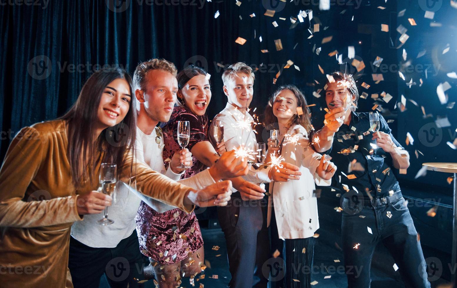 se divertindo com estrelinhas. confete está no ar. grupo de amigos alegres comemorando o ano novo dentro de casa com bebidas nas mãos foto
