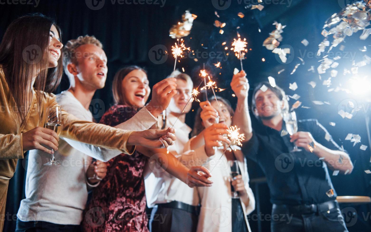 se divertindo com estrelinhas. confete está no ar. grupo de amigos alegres comemorando o ano novo dentro de casa com bebidas nas mãos foto