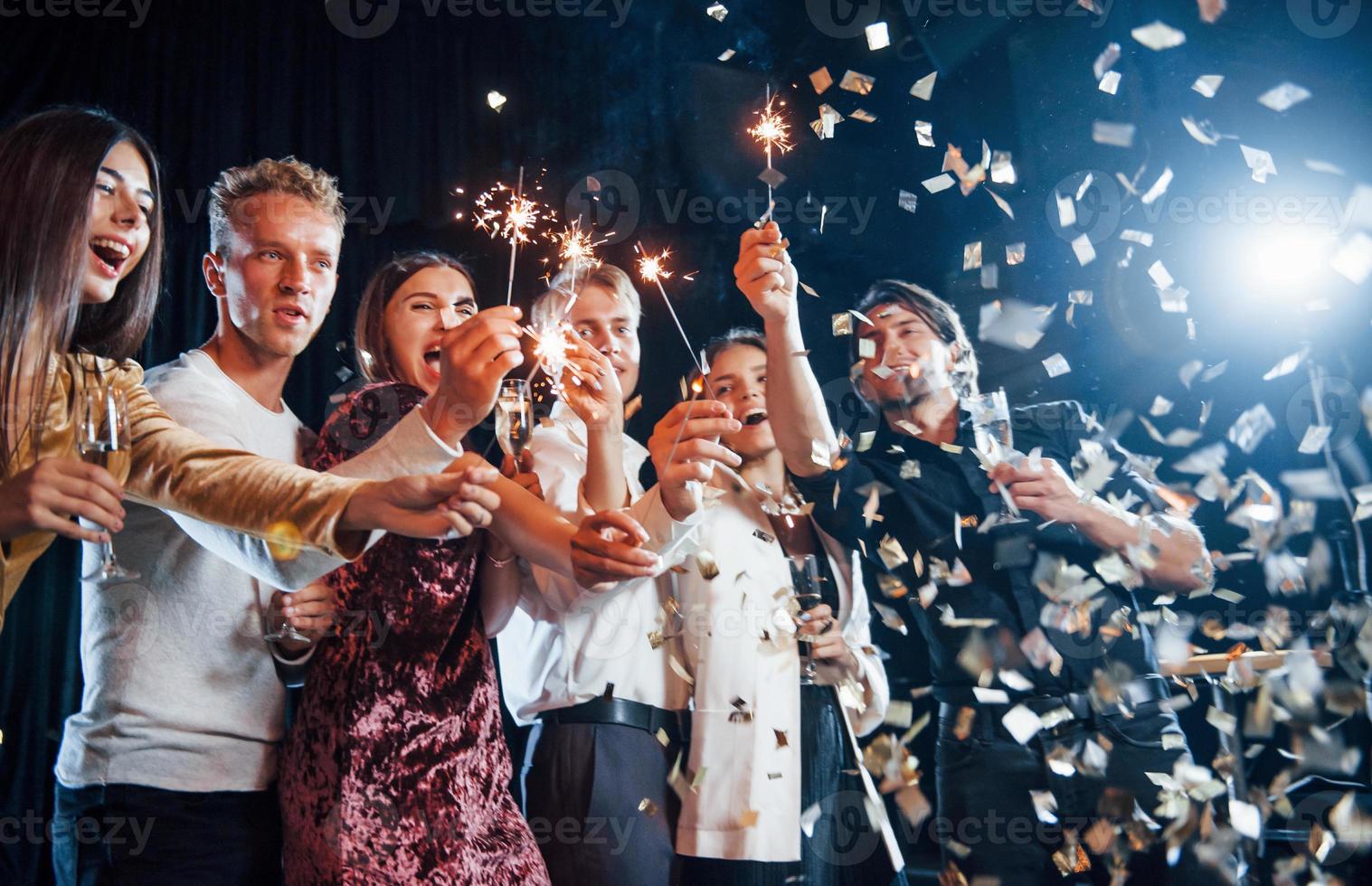 se divertindo com estrelinhas. confete está no ar. grupo de amigos alegres comemorando o ano novo dentro de casa com bebidas nas mãos foto