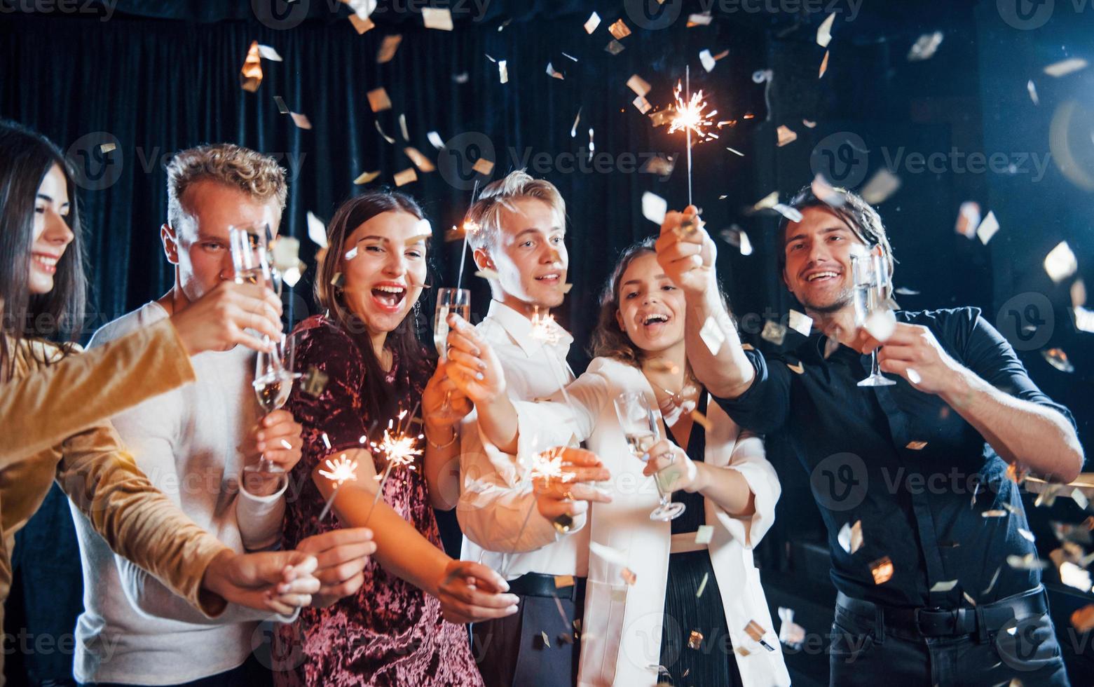 se divertindo com estrelinhas. confete está no ar. grupo de amigos alegres comemorando o ano novo dentro de casa com bebidas nas mãos foto