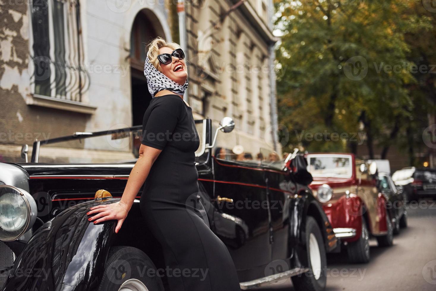 mulher loira de óculos escuros e vestido preto, apoiando-se no velho carro clássico vintage foto