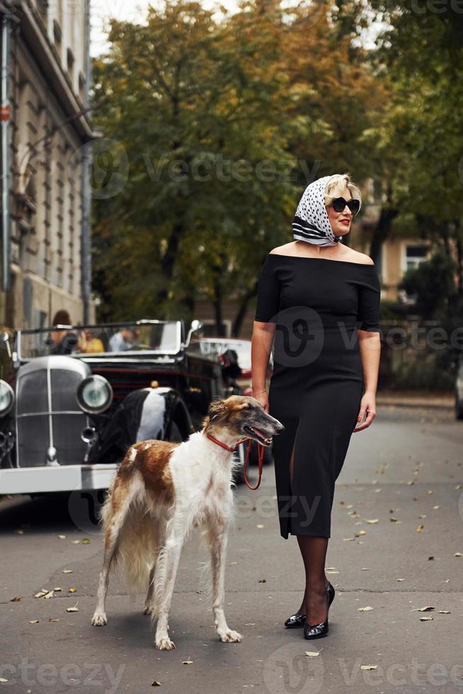 mulher loira de óculos escuros e vestido preto perto do velho carro clássico vintage com seu cachorro foto