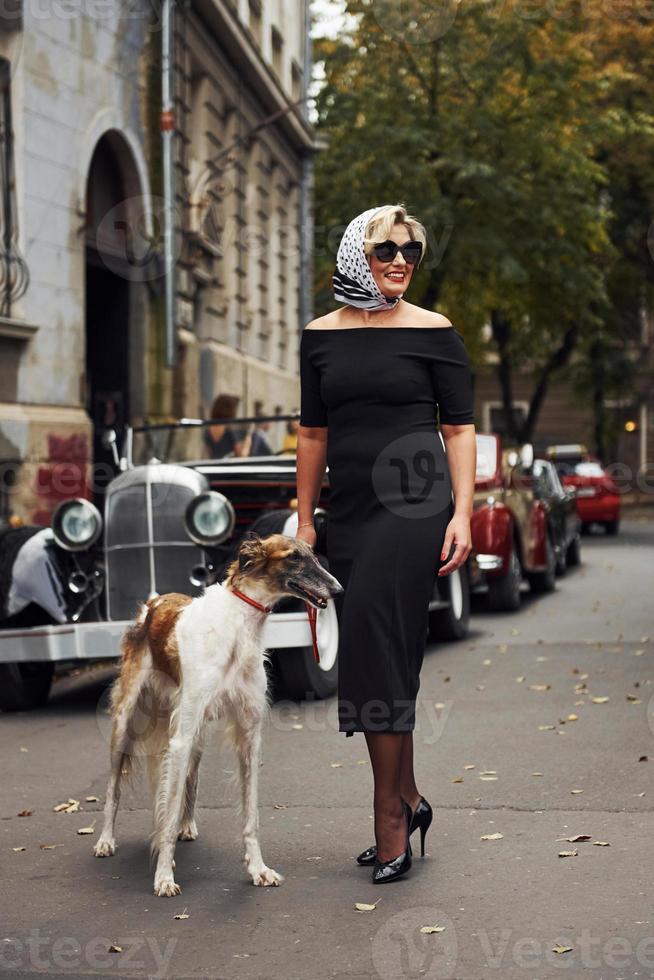 mulher loira de óculos escuros e vestido preto perto do velho carro clássico vintage com seu cachorro foto